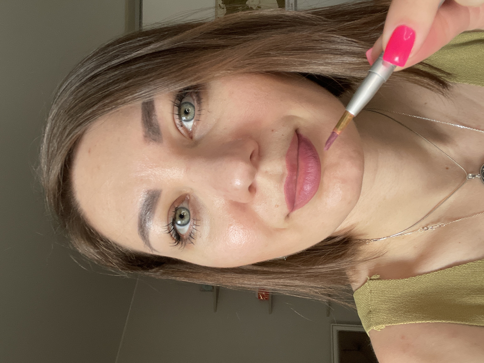 A woman with medium-length brown hair and green eyes is applying pink lipstick with a small brush to her lips. She is wearing a green top and a silver necklace. Her fingernails are painted bright pink. The background is blurred and appears to be an indoor setting. There are shelves visible in the background, one with some items on it. The image is a close-up shot focusing on the woman's face and upper body. It is not possible to determine the specific location in the city of Blagoveshchensk, Russia.