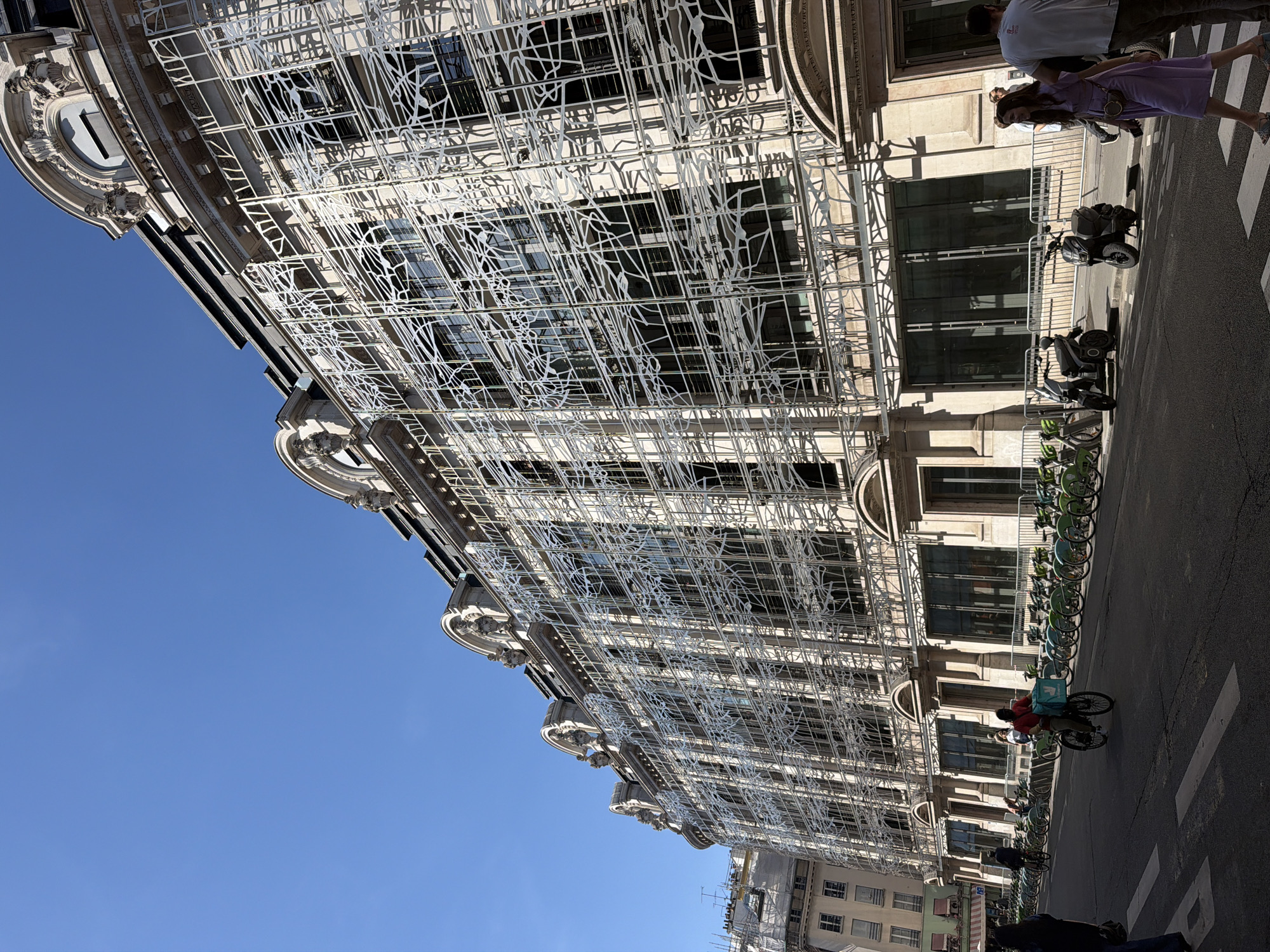 A daytime shot, angled upward, showcases a section of an ornate building in Paris, France. The building's facade is adorned with an intricate, white, metal latticework pattern that obscures much of the windows. Below, on the street, are rows of parked electric scooters and bicycles. People can be seen walking on the sidewalk, and a cyclist wearing a delivery uniform rides by. The sky is a clear, bright blue.