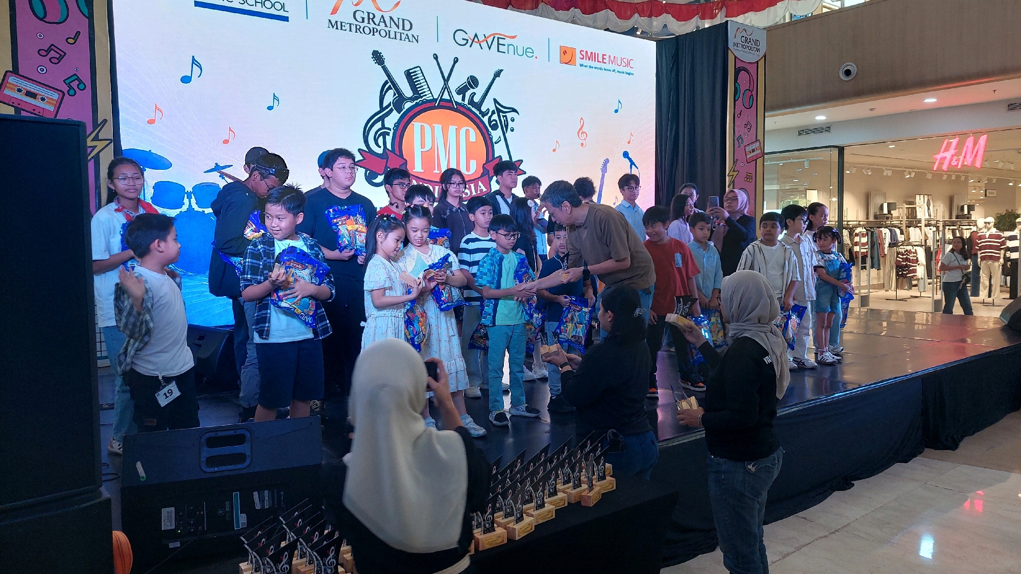 This is a wide shot of an indoor awards ceremony event taking place on a raised stage within a mall. In the background, a large LED screen displays graphics related to a music competition, including logos for "PMC INDONESIA", "GRAND METROPOLITAN", "GAVENUE", and "SMILE MUSIC", as well as musical notes and illustrations of instruments. The backdrop also features a red and white striped banner.
On the stage, a group of approximately twenty children, ranging in age from about 6 to 16 years old, stand in rows. They are holding awards or gift bags. Some of the children are smiling and looking towards the front of the stage, while others are interacting with adults.
In the center of the stage, an adult male in a brown t-shirt is handing out awards to several children. To the right of this interaction, several more children are lined up. In the foreground on the stage, two adult females are also present. One, wearing a grey hijab, is holding a phone and appears to be taking a photo, while the other, also in a hijab, is actively participating in the event, potentially handing out items.
In front of the stage, there is a table with numerous trophies arranged in rows. To the left of the stage, large black speakers are visible. In the far background, through large glass windows, a retail store with clothing displays, identified by an "H&M" sign, is clearly visible, indicating the event is taking place within a commercial mall environment. The floor is tiled. The overall scene suggests a celebratory occasion for young musicians in Bekasi, Indonesia.