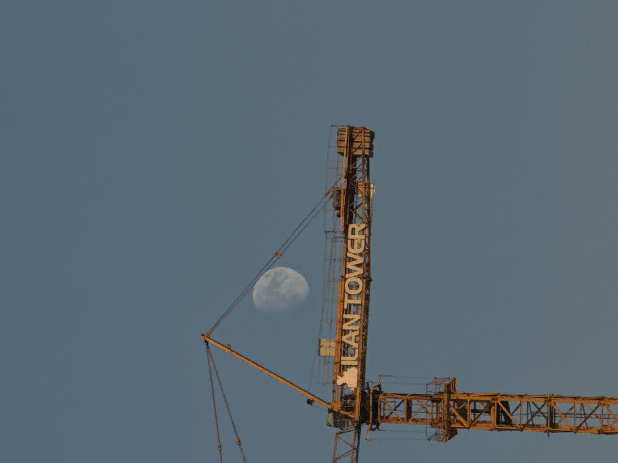 An orange construction crane with the text "ILCAN TOWER" written on its boom is silhouetted against a clear blue sky. A partially visible moon is positioned behind the crane's boom. The crane appears to be in the process of construction, with its tower structure and lifting arm visible. The scene is likely located in the city of Aktau, Kazakhstan, given the context provided.