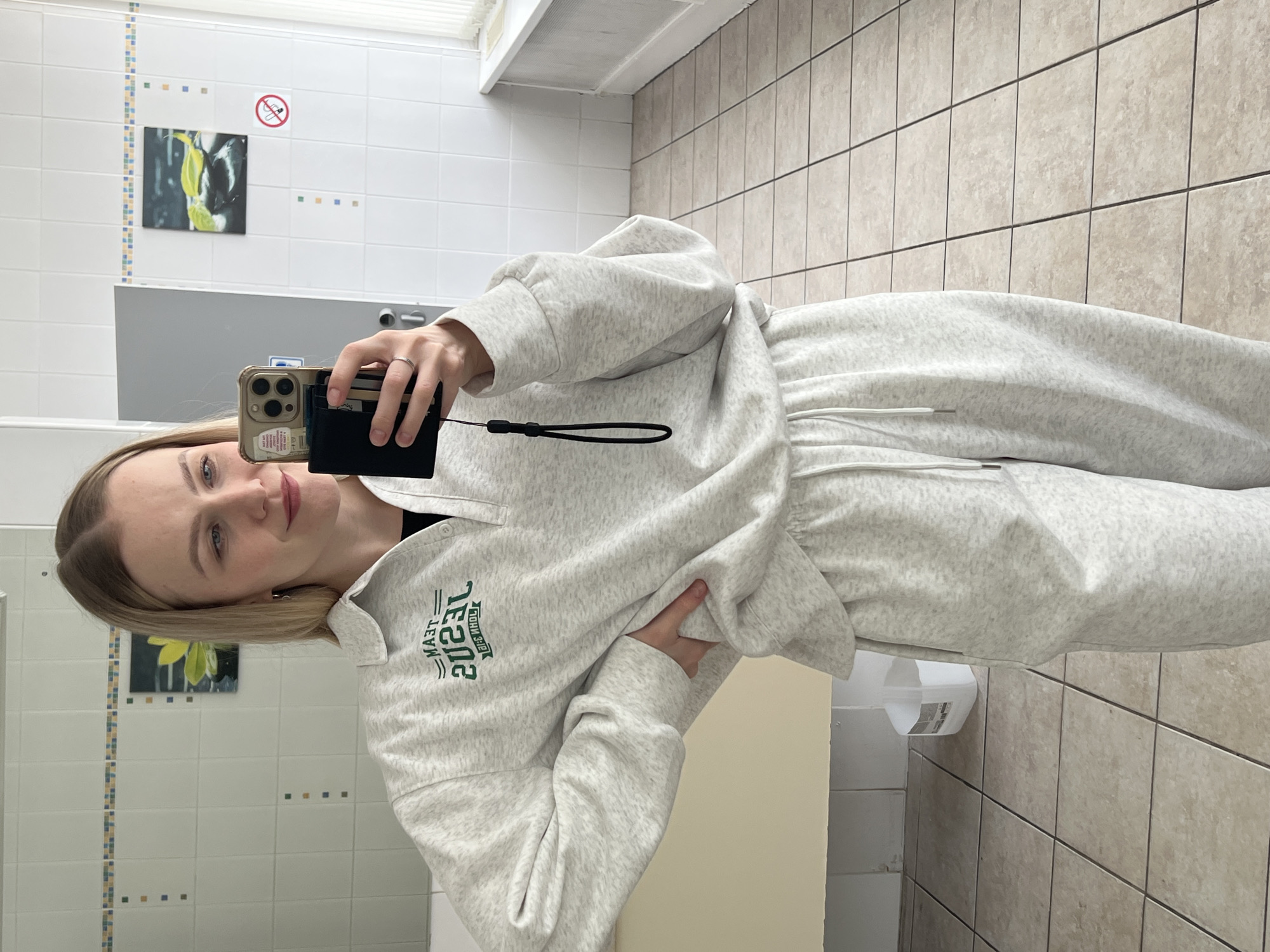 A young woman with blonde hair and light-colored eyes is captured in a mirror selfie, standing in what appears to be a restroom or changing area. She wears a relaxed-fit, light gray heathered tracksuit, comprising a sweatshirt with a collar, partial button placket, and drawstring sweatpants. On the front of her sweatshirt, the green text "TEAM JESUS" and "JOHN 3:16" is visible, reversed due to the mirror reflection.
She holds a gold-colored smartphone, likely an iPhone, in her left hand, which also clutches a black cardholder or wallet with a black wrist strap. A ring is visible on her left ring finger. Her right hand is positioned on her hip, slightly pulling at the sweatshirt. She has a subtle smile on her face.
The background features tiled walls. The upper section has white square tiles with a faint vertical decorative strip of small blue, yellow, and orange tiles. The lower section consists of larger, light beige square tiles. Reflected on the wall behind her in the mirror is a small picture depicting a yellow flower or plant, and a red circular "No Smoking" sign. A white hand dryer is visible in the lower right portion of the mirrored scene. The indoor lighting appears bright and uniform.