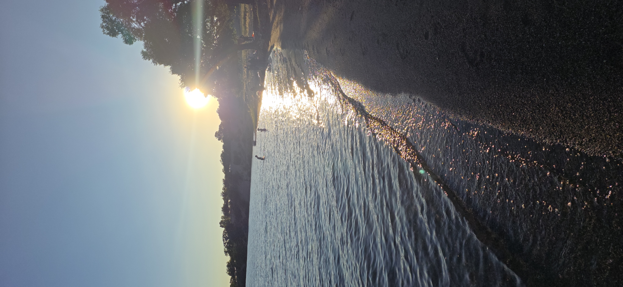 The image depicts a serene sunset scene at a beach on the shore of Issyk-Kul Lake in Kyrgyzstan. The sun, low in the sky, casts a golden glow across the water, creating a shimmering path of light. Gentle waves lap at the sandy shore, where a few figures are visible. Two individuals are wading in the shallow water, appearing to be children. Further down the beach, a group of people are gathered, some seated and others standing near a large tree. The background shows a line of trees and hills under a clear blue sky. The overall atmosphere is tranquil and peaceful, suggesting a warm summer evening.