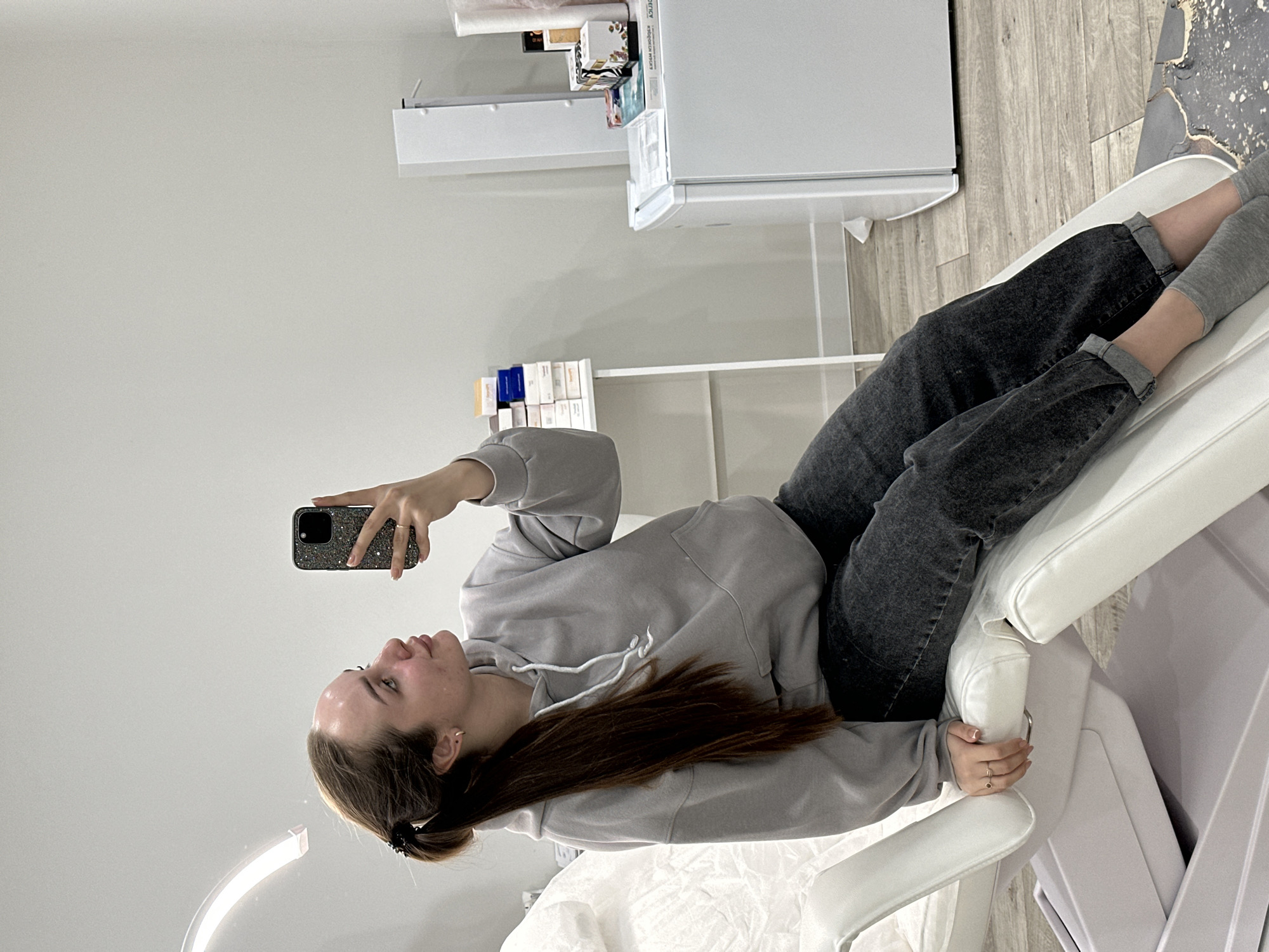 A woman is lying on a white medical examination table, holding a smartphone to take a selfie. She is wearing a grey hoodie and black jeans with grey socks. The room appears to be a medical or beauty treatment space. In the background, there are white cabinets, shelves with boxes, and laminate flooring. The location is likely Chaikovsky, Russia.