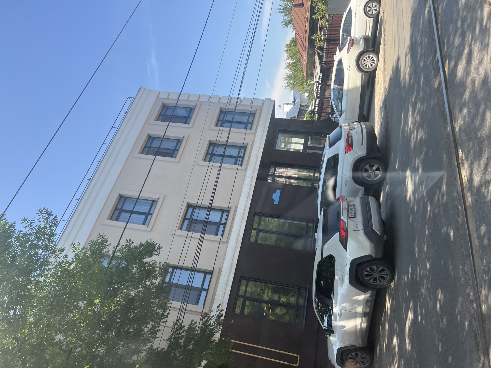 A white Toyota RAV4 is parked on the street in front of a modern building. The building is multi-story and has a light-colored facade with dark lower levels. There are several windows on the building, reflecting the sky and trees. Other cars are also parked along the street. The scene appears to be in a city, with trees and power lines visible. The license plate of the Toyota RAV4 is partially visible, showing Russian characters. Based on the presence of Russian license plates and architectural style, the location is likely in Ulyanovsk, Russia.