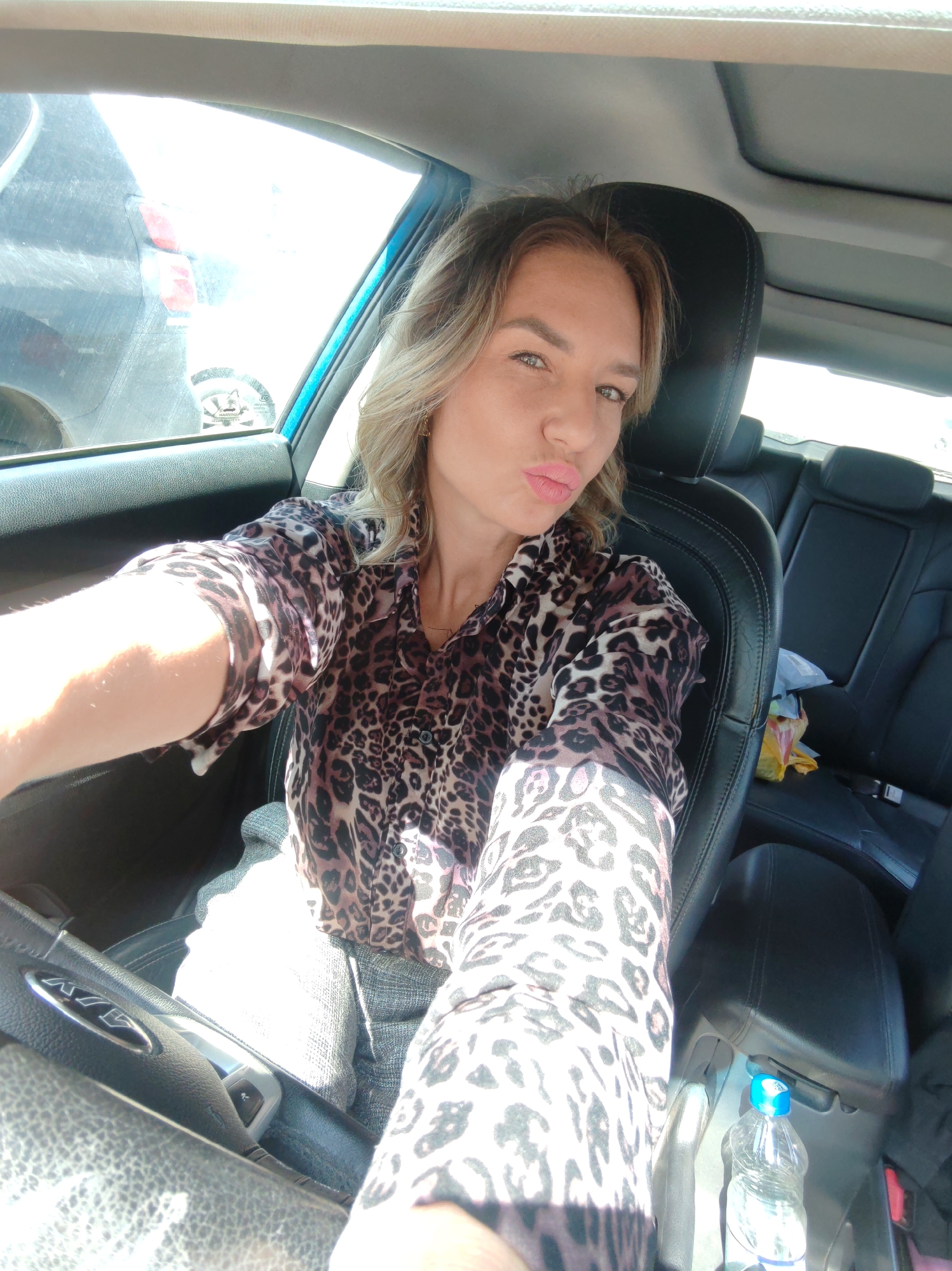 The image is a selfie of a woman taken from inside a car, likely in the driver's seat, set in Petropavlovsk-Kamchatsky, Russia. The woman, with light brown, wavy hair, is looking directly at the camera with a playful "kissing face" expression, accentuated by pink lipstick. She is wearing a button-up shirt with a prominent brown and black leopard print pattern, and light-colored pants or shorts are visible below.
The car's interior features black leather seats, including the headrest behind her and the rear seats, where a white and a yellow plastic bag are placed. A sunroof is visible in the ceiling of the car. The steering wheel, partially visible on the left, displays a "KIA" logo. Bright sunlight streams through the driver's side window, creating glare on the window and illuminating her left arm and shoulder. Through this window, the rear of a dark-colored vehicle with red taillights is partially visible, indicating an outdoor parking location. A blurry warning sticker, with text like "WARNING" and "SECURITY" partially legible, is affixed to the driver's window. A clear plastic water bottle with a blue cap is situated in the center console. The bright lighting suggests it is daytime with sunny weather. The woman's pose conveys a confident or lighthearted mood.