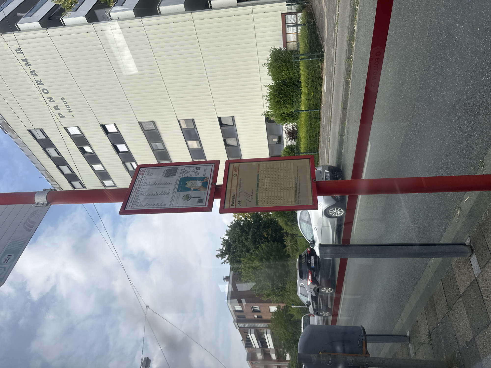 A street-level, eye-level medium shot shows a bus stop in Krefeld, Germany, with a large apartment building in the background. The bus stop features a red pole and two timetable displays. The larger display, on the left, contains general information and a QR code. The smaller display, on the right, shows the timetable for line 044, heading towards "Richtung Hüls Bethel." A portion of the building's facade is visible to the right of the bus stop, displaying the word "PANORAMA" and "- Haus". In the middle ground, several cars are parked or driving on the street, including a black SUV and a silver hatchback. The foreground consists of a sidewalk and the road, separated by a reflective red barrier. The sky is overcast with clouds.