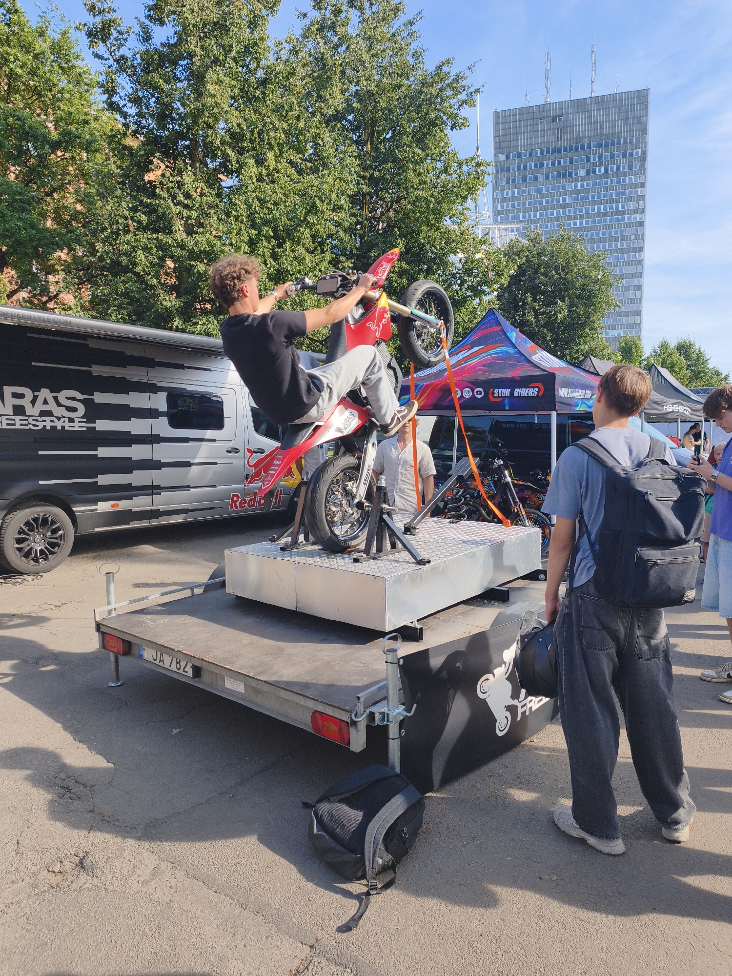 The image shows a young man performing a motorcycle stunt on a raised platform in what appears to be an outdoor event or demonstration. The motorcycle is red and silver, and the rider is in mid-air, with the front wheel of the motorcycle lifted. He is wearing a black t-shirt and light-colored pants. Behind him, there is a large silver van with "ARAS FREESTYLE" written on its side. A blue and red tent with the text "STUNT RIDERS" and a website address is also visible. In the background, a tall, modern glass-fronted building stands out against a clear blue sky. To the right of the frame, two young men are observing the stunt. One is wearing a gray t-shirt and a black backpack, while the other is in a purple t-shirt and denim shorts. A trailer with a license plate reading "JA 78Z" is in the foreground. The ground is asphalt, and there are shadows cast by the subjects and objects.
The tall glass building in the background is likely the Latvian Academy of Sciences building, a prominent landmark in Riga, Latvia. This suggests the event is taking place in the vicinity of the Academy of Sciences.