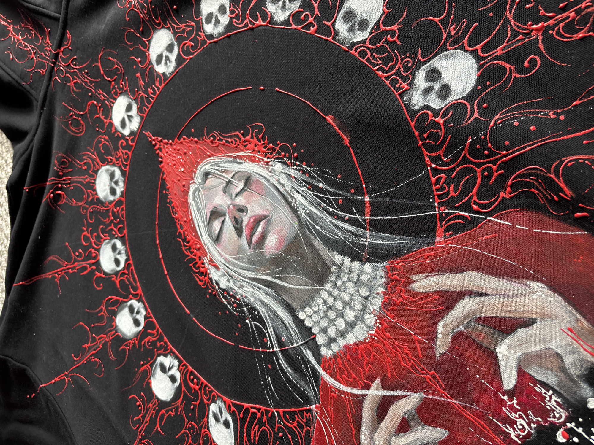 A detailed close-up photograph captures a gothic-inspired artwork, likely painted or screen-printed onto black fabric, possibly a hoodie or garment.
The central subject is a figure, predominantly a woman with pale skin, closed eyes, and flowing white or silver hair. Her lips are painted a striking red. She wears a prominent red, pointed hood or hat, and a necklace or collar composed of numerous white, pearl-like beads. Her hands, with slender, pale fingers, are visible on the right side of the composition.
The figure is framed within a large black circular element. Surrounding this circle, and extending across the black fabric, are numerous small, white or grey skulls. These skulls are arranged in a pattern, interspersed with raised, three-dimensional red tendrils or vine-like patterns that appear to be made with puff paint or a similar textured medium. Additional red and white paint splatters are scattered across the artwork, adding to its dynamic and expressive quality.
The overall scene conveys a dramatic and macabre aesthetic, with the figure appearing in a state of serene repose, sleep, or death, emphasized by the surrounding skulls and dark coloration. The artwork itself contains no specific location cues or visible text, but the photograph originates from Krasnoyarsk, Russia. The time of day or specific weather is not discernible from the artwork.