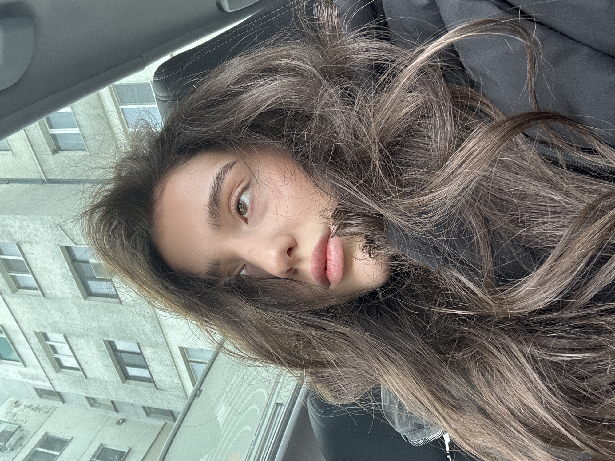 A young woman is photographed from a close-up, low-angle perspective, giving the impression she is reclining and looking down at the camera. She has long, wavy, brown hair that frames her face. Her skin appears clear, and her lips are full and slightly parted. Her eyes are light brown. She is wearing a dark jacket. The background reveals a building with several windows, suggesting an urban setting. The photo seems to be taken from inside a car. The city location is Unknown.