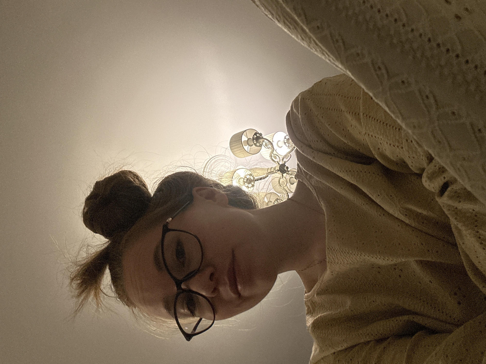 A young woman is captured in a low-angle shot, looking down towards the camera from an overhead perspective. She has light-colored hair gathered into a messy bun on top of her head and wears black-framed glasses. Her expression is neutral or slightly pensive as she gazes downward. She is dressed in a light-colored, possibly cream or beige, long-sleeved top featuring a textured or patterned fabric, possibly knitted or crocheted.
Behind her head and slightly above, a decorative chandelier with multiple arms and shaded light fixtures is visible, emitting a bright glow. This artificial light illuminates the plain, light-colored ceiling or wall that forms the background. A section of a textured, light-colored blanket or textile hangs into the frame from the upper right, partially obscuring the background. The scene depicts an indoor setting, likely a home. No specific activity is taking place beyond the subject looking at the camera. There are no discernible location cues, visible text, or clear indicators of the time of day or weather.