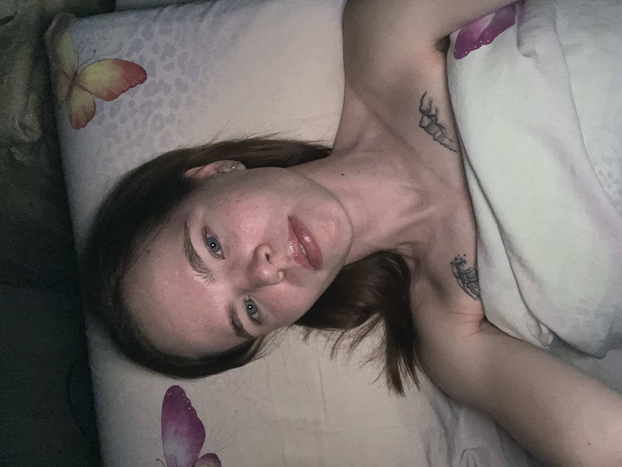 A woman lies on a bed with a butterfly-patterned pillow and bedding. She has brown hair, blue eyes, and two visible tattoos on her left side. One tattoo is on her shoulder/neck area, and the other is on her arm. No information in the image indicates this scene is in Chaikovsky, Russia.