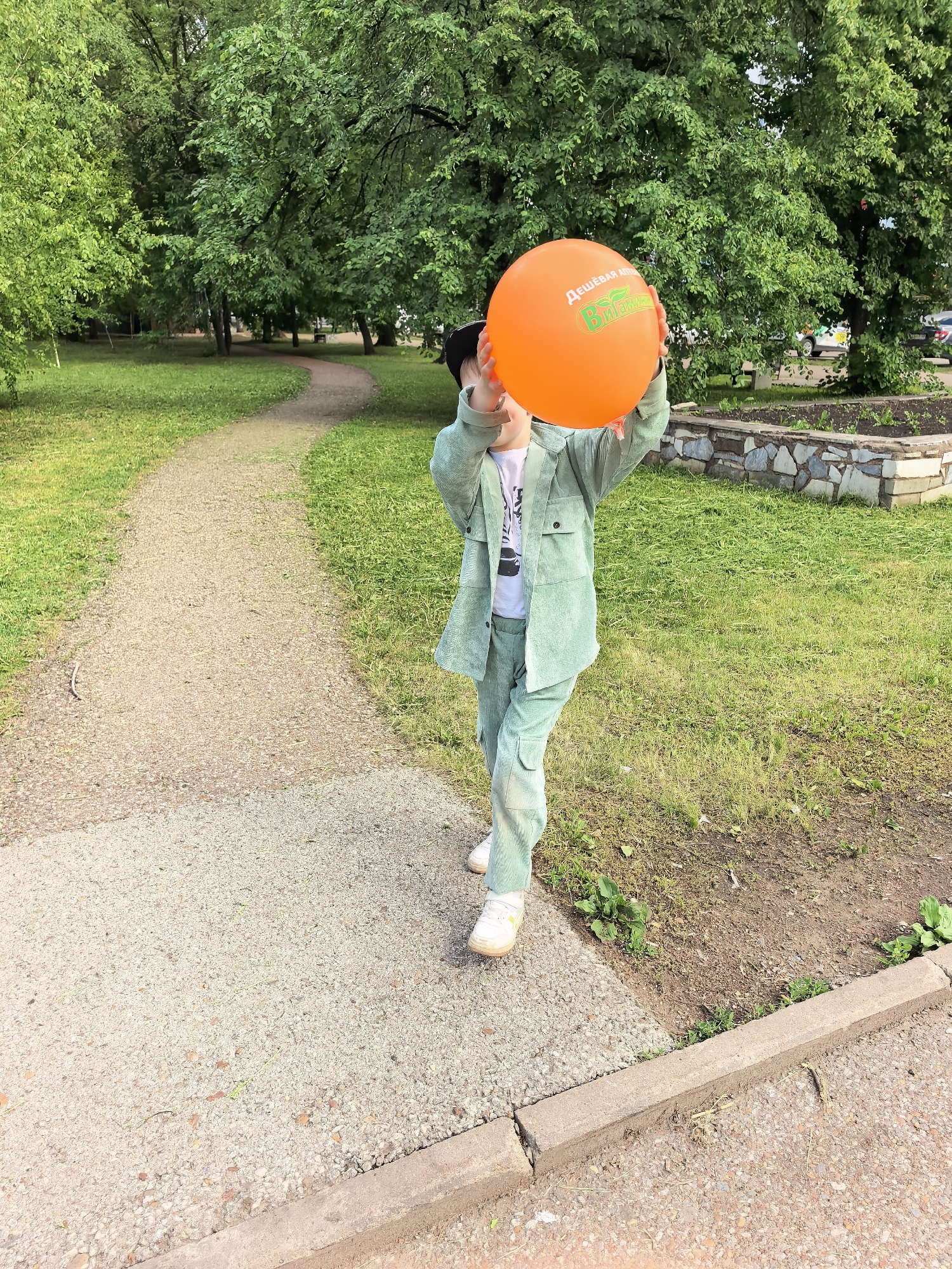 The image captures a child holding an orange balloon outdoors, likely in a park setting. The child is wearing a light green corduroy suit and white sneakers. The balloon has text in Russian, indicating a possible advertisement or promotion. The background features a gravel path, grassy areas, trees, and a stone-edged flower bed. The location is identified as Sterlitamak, Russia.
