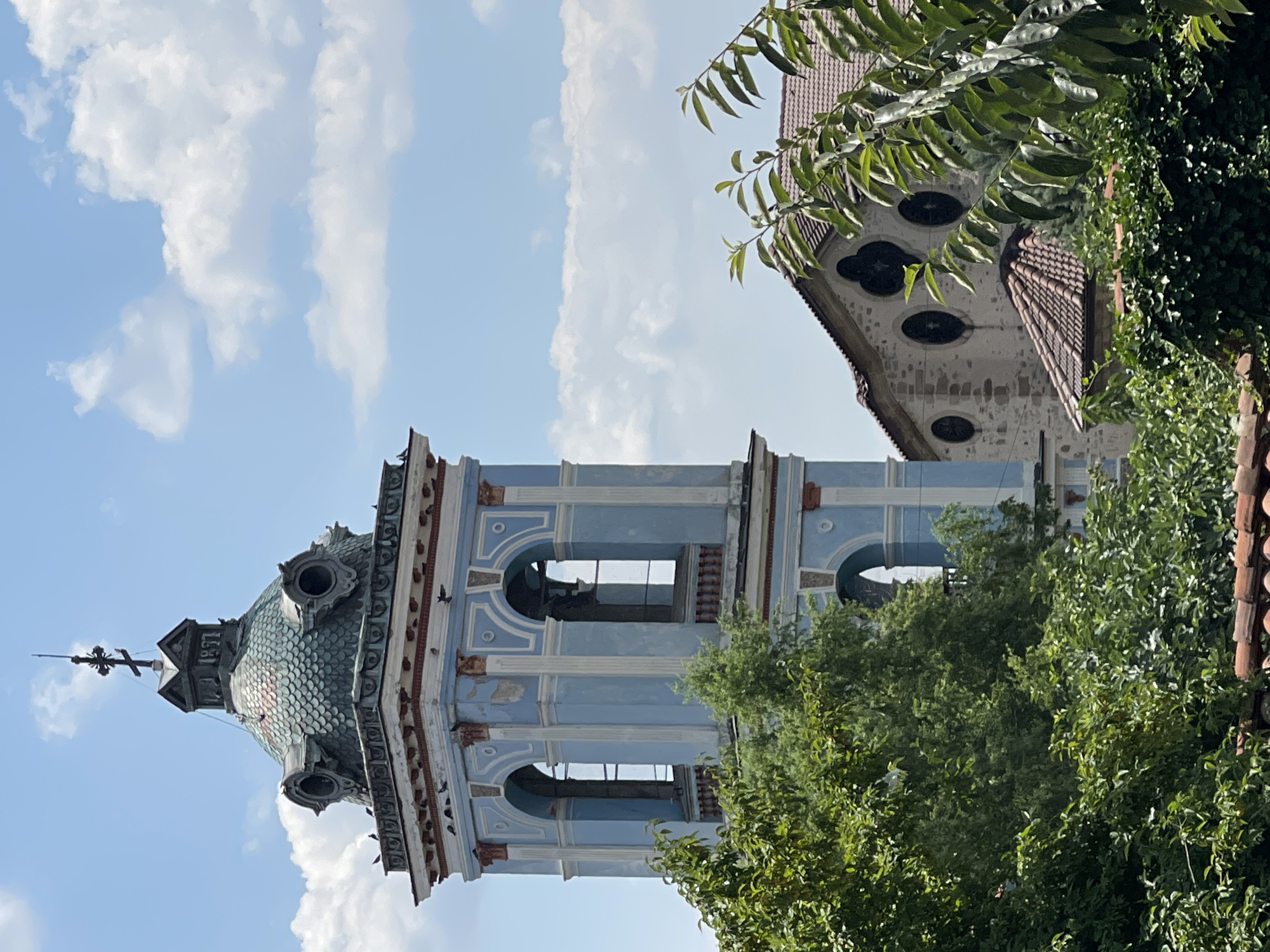 The image shows a blue bell tower with a green tiled roof, topped with a cross, standing against a blue sky with scattered clouds. The bell tower has arched openings with visible bells inside. Below the bell tower, a portion of a stone building with a red tiled roof is visible. Lush green trees and foliage partially obscure the lower parts of the bell tower and the adjacent building. The scene depicts a historical architectural structure, likely a church or monastery, in Karlovo, Bulgaria. There are no people or animals visible in the frame.