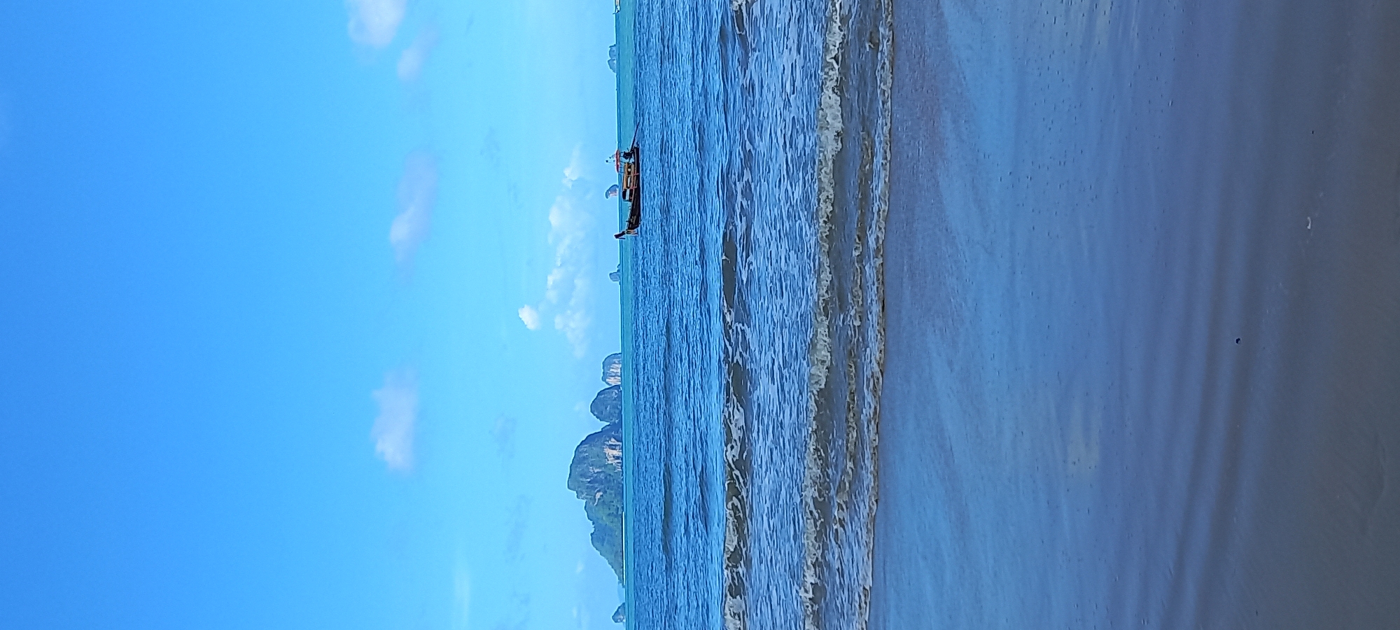 The image depicts a coastal scene near Ban Klong Haeng, Thailand. A longtail boat is visible in the water. The shoreline exhibits the transition from ocean waves to a sandy beach. The sky is mostly clear with some scattered clouds. In the distance, rocky island formations emerge from the sea.