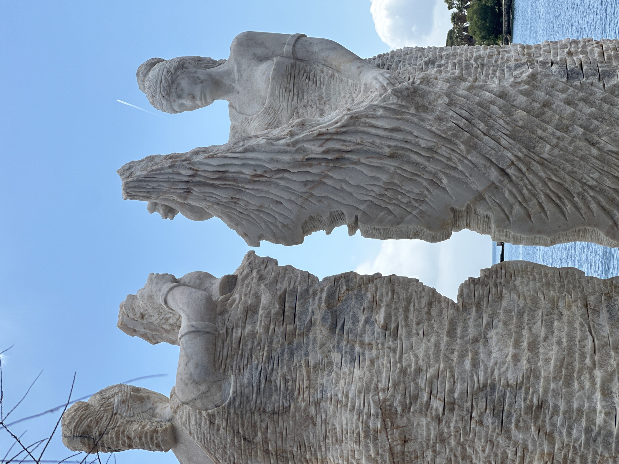 A low-angle, rotated image depicts a monumental marble sculpture of three female figures against a backdrop of clear blue sky, a contrail, trees, and water. The sculpture is carved with visible vertical striations and layered horizontal detail in the draped clothing. The top figure's head is adorned, and she wears a banded garment. The two figures below are less distinct but appear similarly dressed. The scene suggests an outdoor setting, possibly a coastal park or public art space in Datça, Turkey.