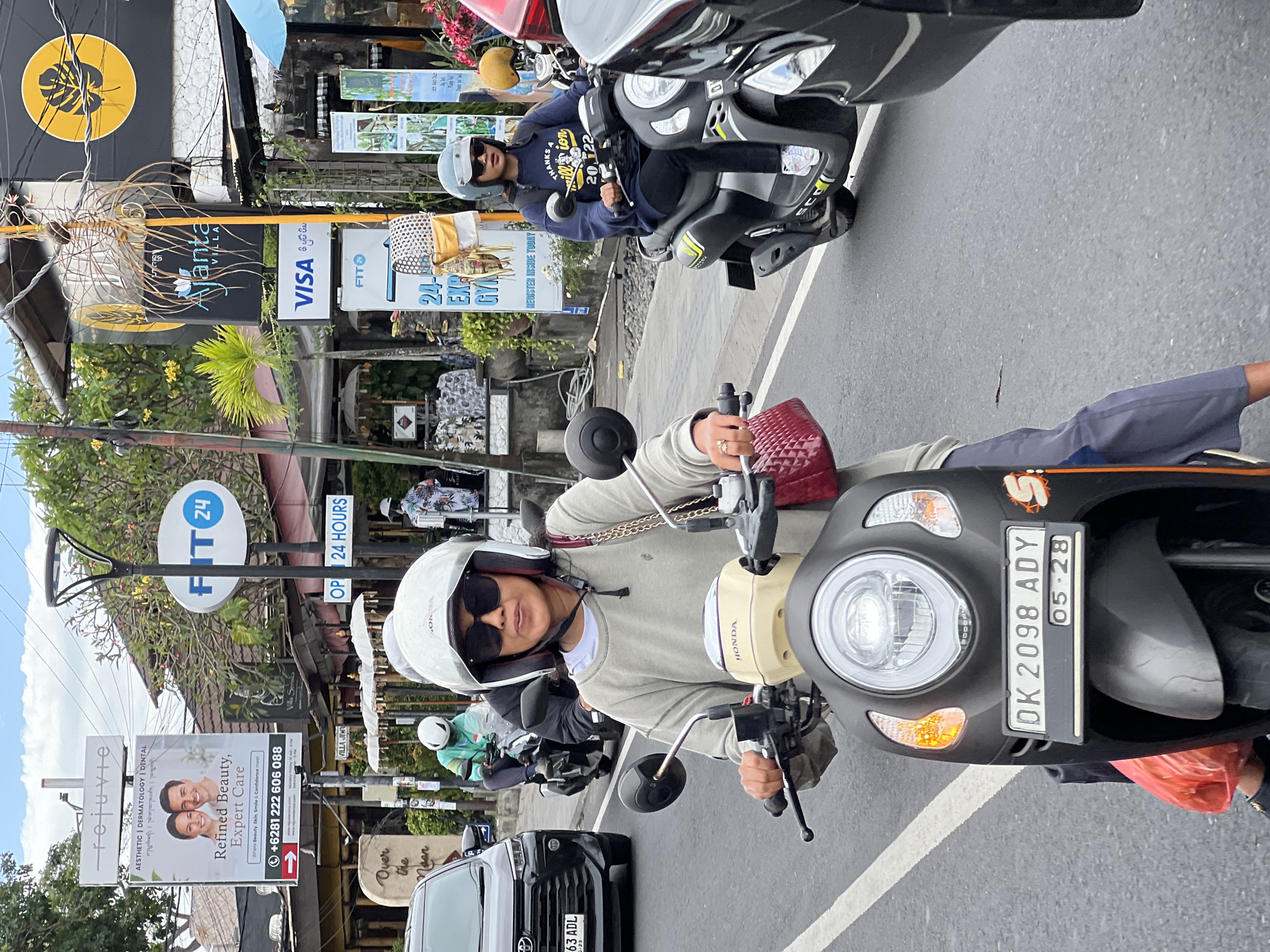 The image captures a street scene in Denpasar, Indonesia, on a cloudy day. A woman wearing a white helmet and sunglasses is riding a grey and beige Honda scooter in the foreground. She is wearing a light grey long-sleeved shirt and dark grey pants. To her right, another woman on a dark grey scooter wears a navy blue long-sleeved shirt, dark pants, and a white helmet with sunglasses. Behind them, a person in a light blue shirt and white helmet rides a scooter. Several businesses with signage are visible in the background, including "rejuvie aesthetic | dermatology | dental", "FIT 24", and "Ajanta Villa". A black Toyota car is parked on the left side of the road. The street appears to be a busy thoroughfare with traffic.