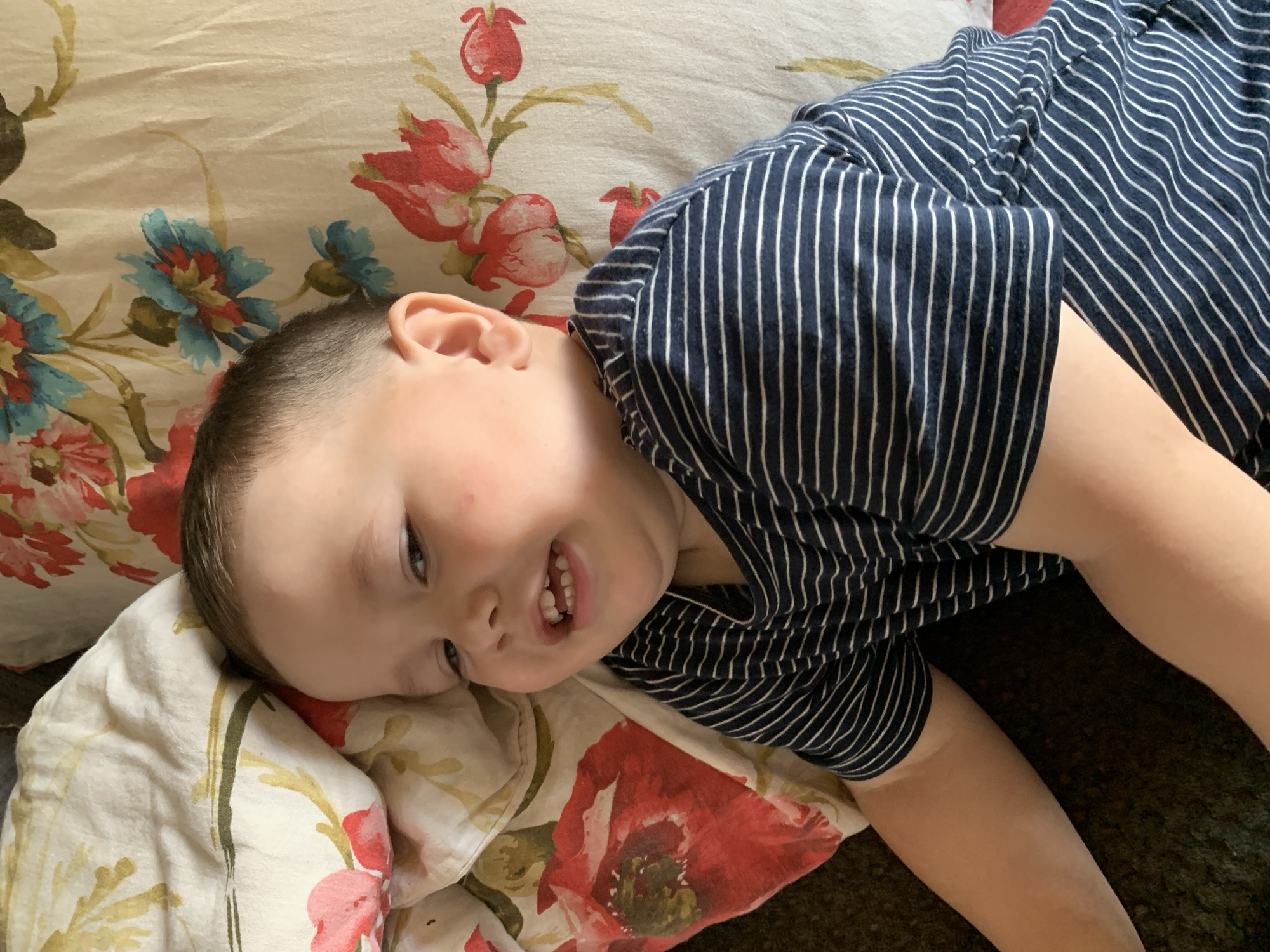 In the city of Фоки, Russia, a young child with light skin and brown hair is lying on a bed with a floral-patterned comforter. The child is wearing a navy blue and white striped t-shirt. The child is looking to the left of the frame, with their mouth slightly open, revealing teeth. The lighting suggests an indoor setting, possibly a bedroom.