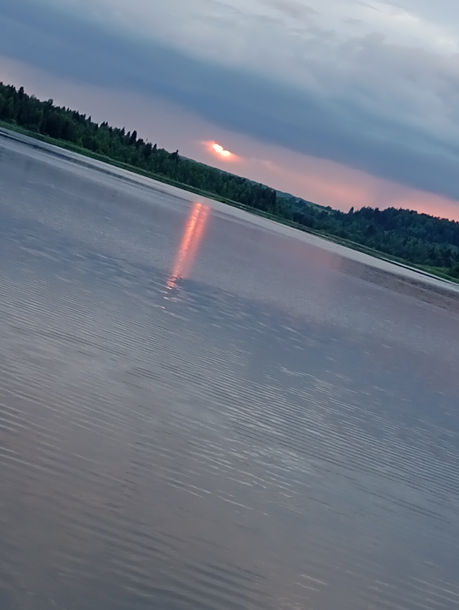The image depicts a body of water, likely a river or lake, with a shoreline of dense green trees. The sky is overcast with dramatic clouds, and a faint pink glow suggests either sunrise or sunset. The water's surface is rippled, and a bright, elongated reflection of the light source is visible on the water. The image is taken from a slightly elevated angle, looking across the water towards the treeline. The location of Рябки, Russia is not directly ascertainable from the visual information provided.