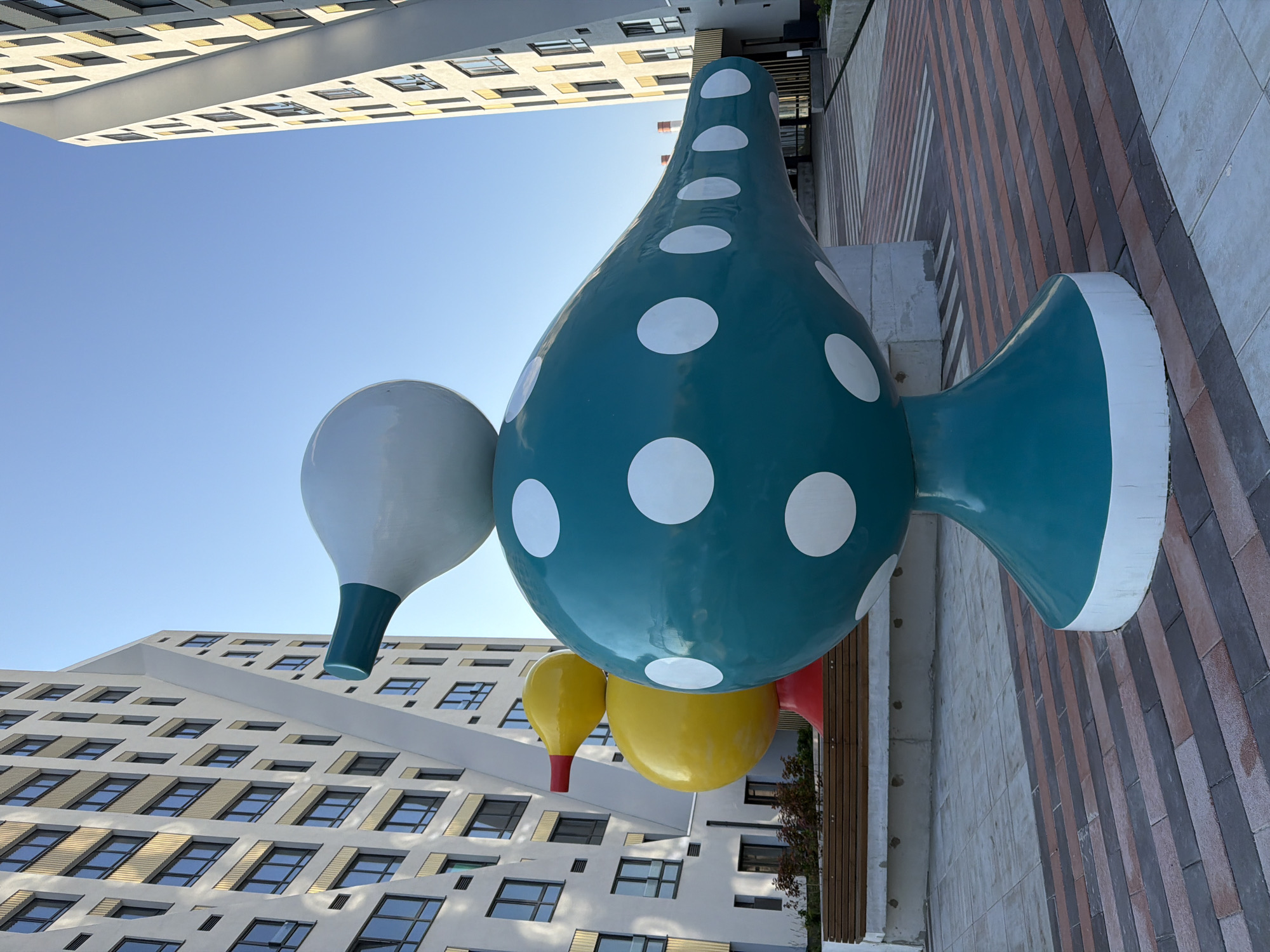 The image depicts a public art installation of abstract, bird-like sculptures in a city setting. The foreground is dominated by a large, teal sculpture with white polka dots, resting on a teal and white base. Behind it, a yellow sculpture with a red beak is visible. These sculptures are situated in a paved outdoor area with patterned brickwork. In the background, modern, multi-story apartment buildings with repeating window patterns rise against a clear blue sky. The architecture suggests a contemporary urban environment. The sculptures are positioned in a courtyard or open space between the buildings, with benches visible in the middle ground. This scene is located in the city of Yekaterinburg, Russia, specifically in the residential complex "Minimal" on Shirokaya Street.