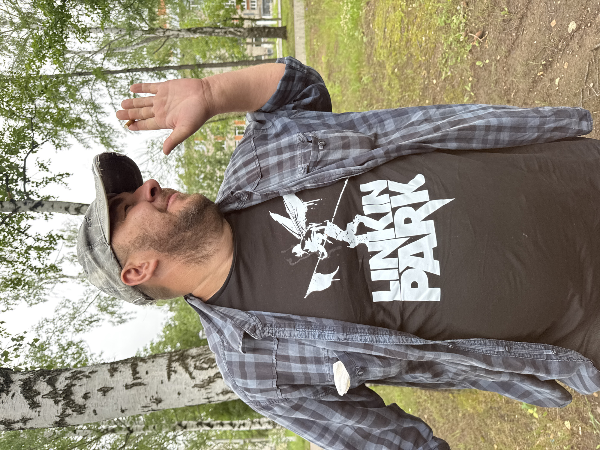 A man wearing a grey camouflage baseball cap, an unbuttoned grey and black plaid shirt over a black t-shirt with a white Linkin Park logo, is standing outdoors in a park with birch trees. He has a beard and is raising his right hand, palm facing forward, as if waving or signaling. A white object, possibly a tissue, is in the chest pocket of his shirt. The background shows grass, dirt, and buildings in the distance, consistent with an urban park environment in Chaikovsky, Russia. The weather appears overcast.