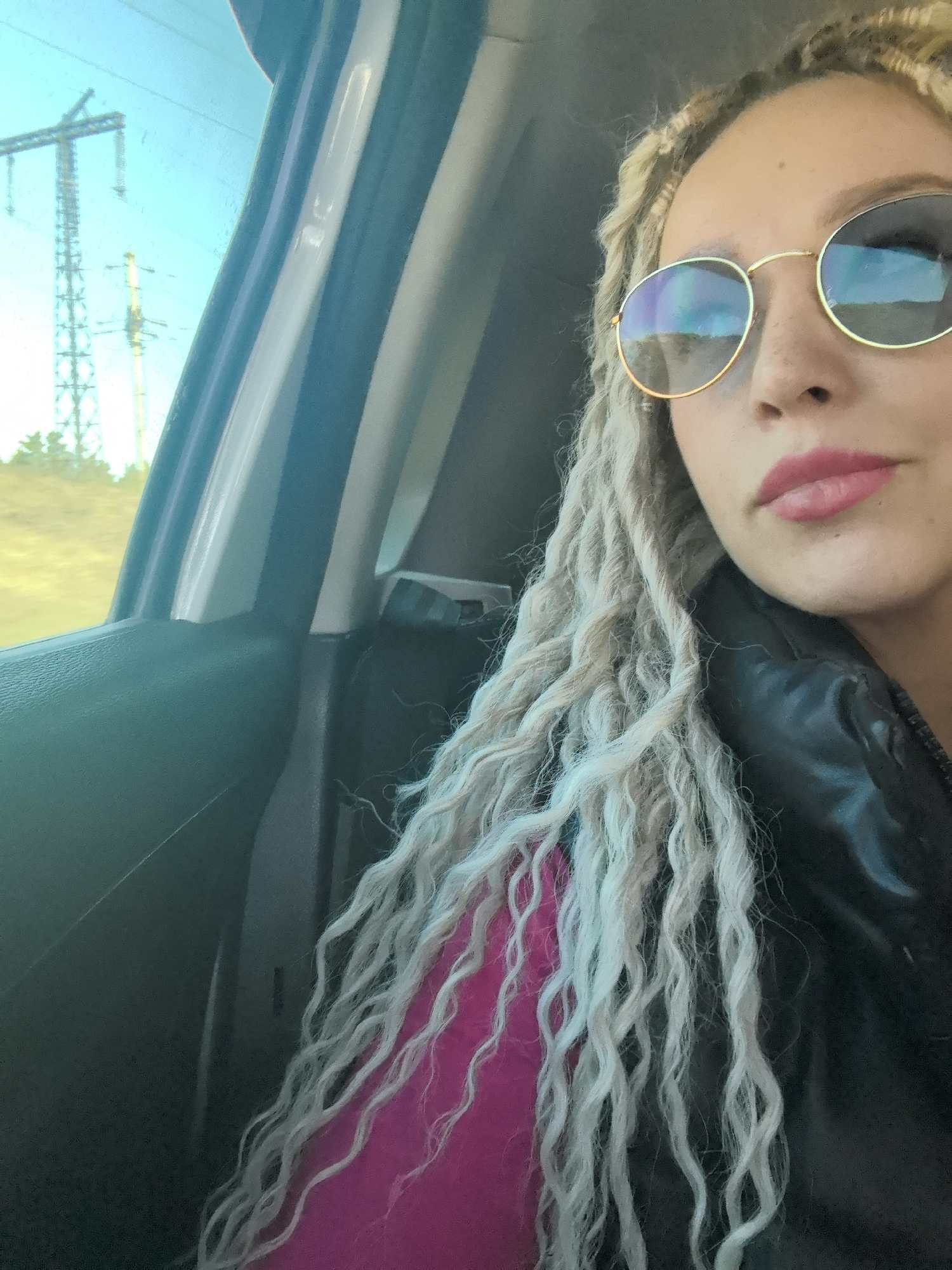 A person with blonde dreadlocks and round sunglasses is inside a car. The person is wearing a black jacket over a pink shirt. Outside the car window, utility poles are visible against a sky. The photo appears to be taken during daytime. Based on the request, the location is specified as Chaikovsky, Russia.