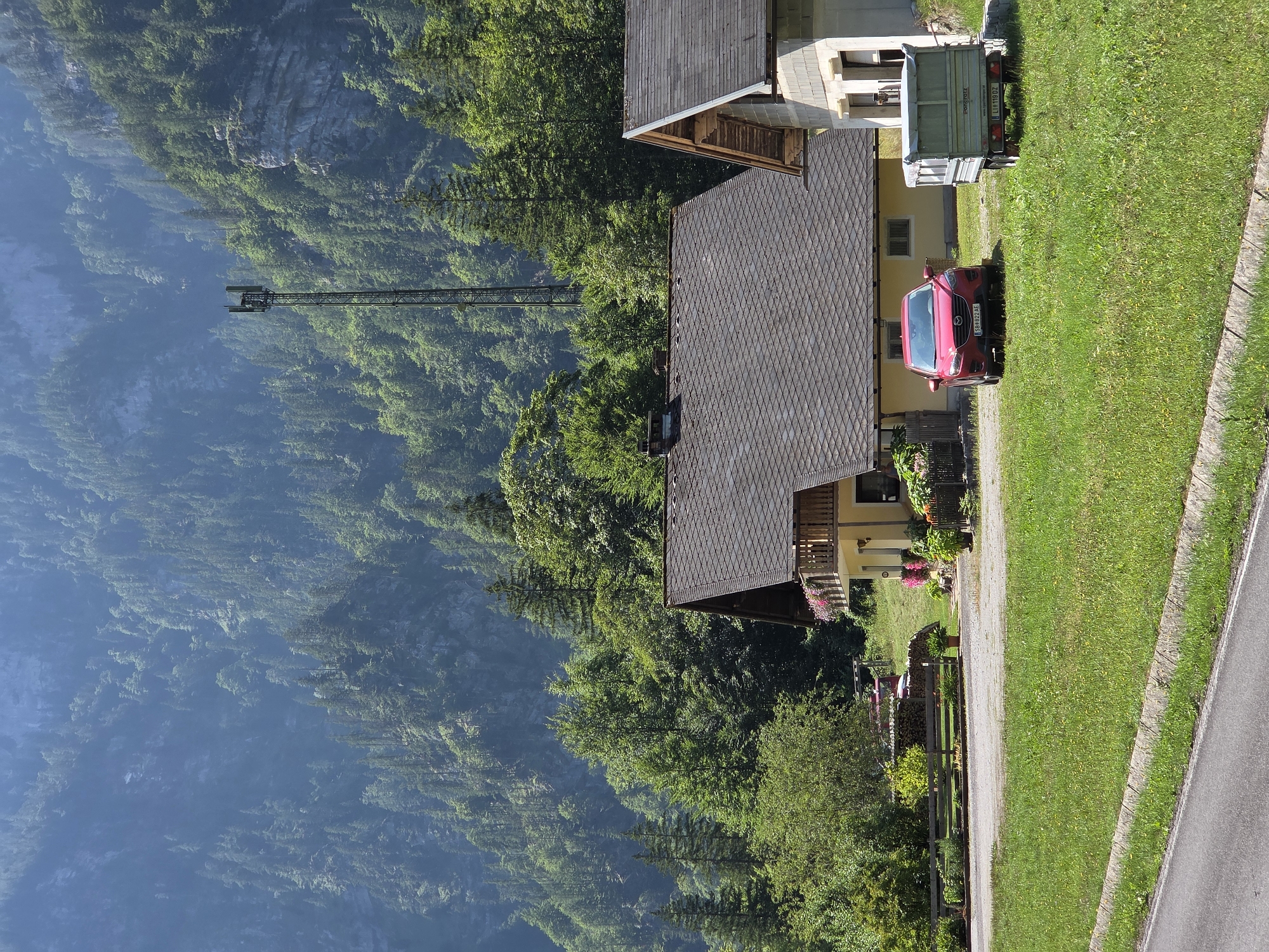 The image depicts a rural scene in Landl, Austria, featuring a cluster of houses nestled at the base of a steep, forested mountain. In the foreground, a paved road runs along the bottom left of the frame, bordered by a grassy verge and a stone curb. A well-maintained lawn stretches across the middle ground, dotted with small yellow wildflowers.
A bright red car, identified by a license plate that appears to read "SB 822 AE," is parked on a gravel area. Beside the car, a small, light-colored utility trailer with the license plate "LI 119 DZ" is hitched to an unseen vehicle or parked on its own.
Dominating the middle ground are two houses. The larger house on the left has a dark, pitched roof with diamond-shaped tiles, a yellow facade, and a wooden balcony adorned with pink flowers. The smaller house on the right is partially visible, with a similar pitched roof and a concrete block facade.
Behind the houses, a wooden fence runs horizontally, separating the lawn from a sloped area with more vegetation and a stack of firewood. Beyond this, the landscape rises sharply, covered in a dense forest of coniferous and deciduous trees. The mountain slopes are steep and rugged, with rocky outcrops visible in places. In the background, a tall, dark metal telecommunications tower stands prominently against the green foliage. The sky above is clear and bright, suggesting a sunny day. The overall impression is one of a serene, mountainous village setting.