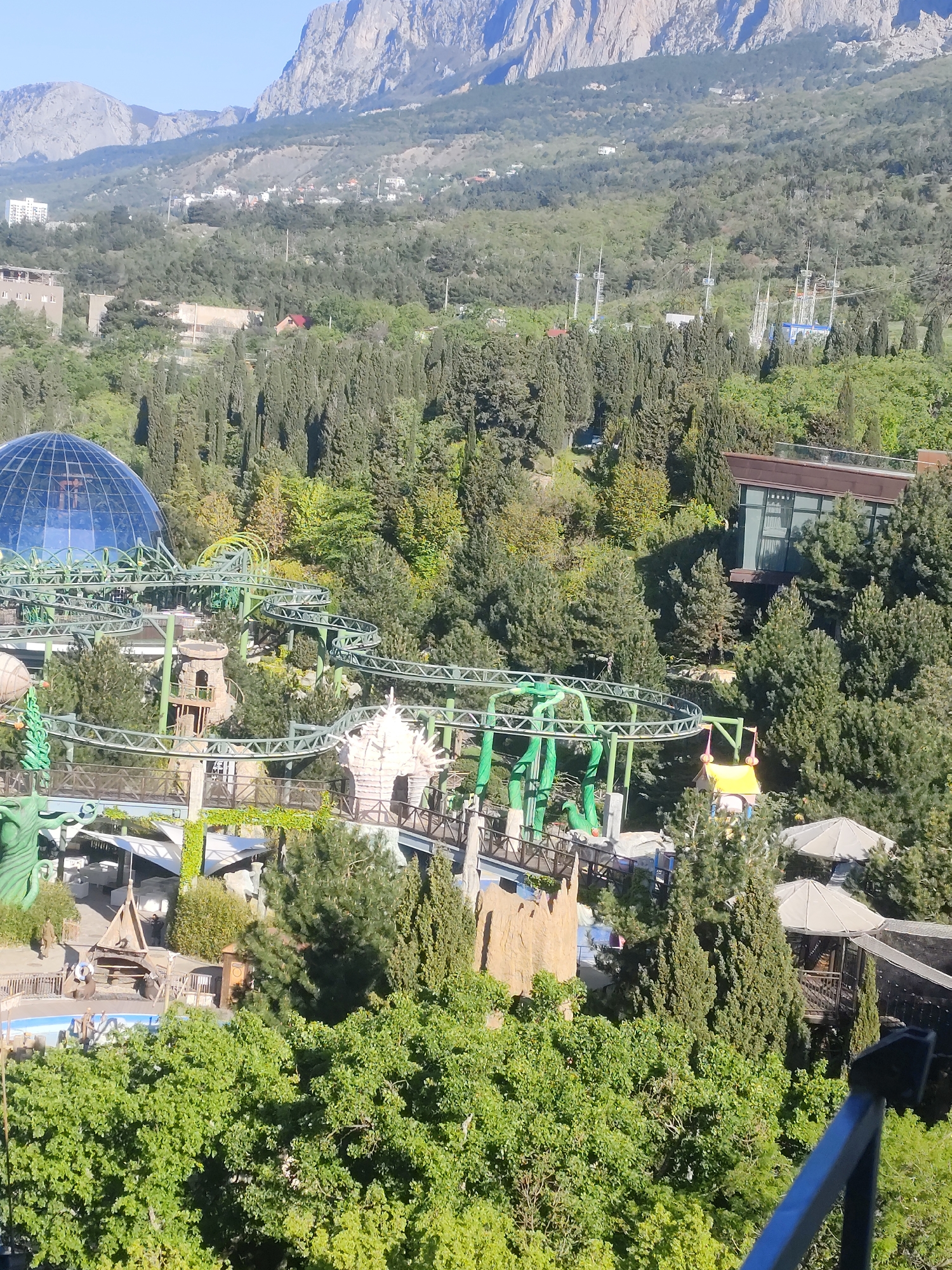 The image captures a high-angle view of a recreational area, possibly a theme park, nestled amidst lush greenery. A notable feature is a winding roller coaster structure, painted in a combination of light gray and vibrant green, that weaves through trees and various architectural elements.
In the background, a partially visible geodesic dome with a blue hue adds a distinctive architectural element. Beyond the immediate park area, a mountainous landscape looms, with residential buildings and structures scattered across the lower slopes.
The image's composition, combined with the presence of recreational features, suggests a location situated in a city or town in close proximity to a natural landscape.