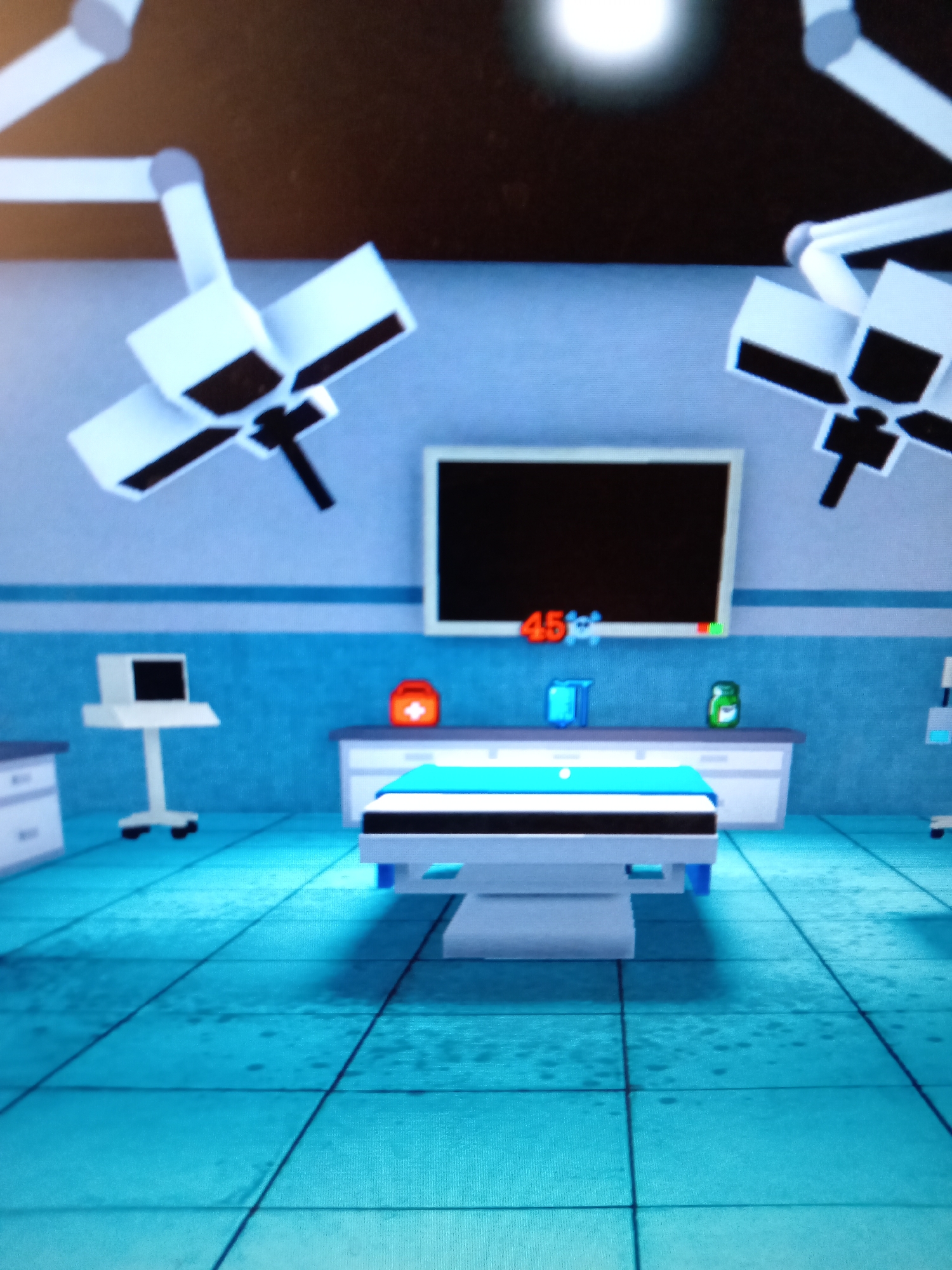 The image depicts a digital rendering of an operating room. The room is painted in light blue with a tiled floor in the same color. Two large, ceiling-mounted surgical lights are visible. In the center of the room, an operating table with a light blue mattress is positioned. Behind the table, a counter with medical supplies, including a red first-aid kit, a blue container, and a green bottle, is placed against the wall. A large monitor screen is mounted on the wall above the counter, displaying the number "45%" in red. To the left of the operating table, a medical monitor on a stand is visible. The overall impression is a sterile, high-tech environment, typical of a modern surgical facility. The location in the city of Rudny, Kazakhstan, is not discernable from the image itself, as it appears to be a stylized or virtual representation.