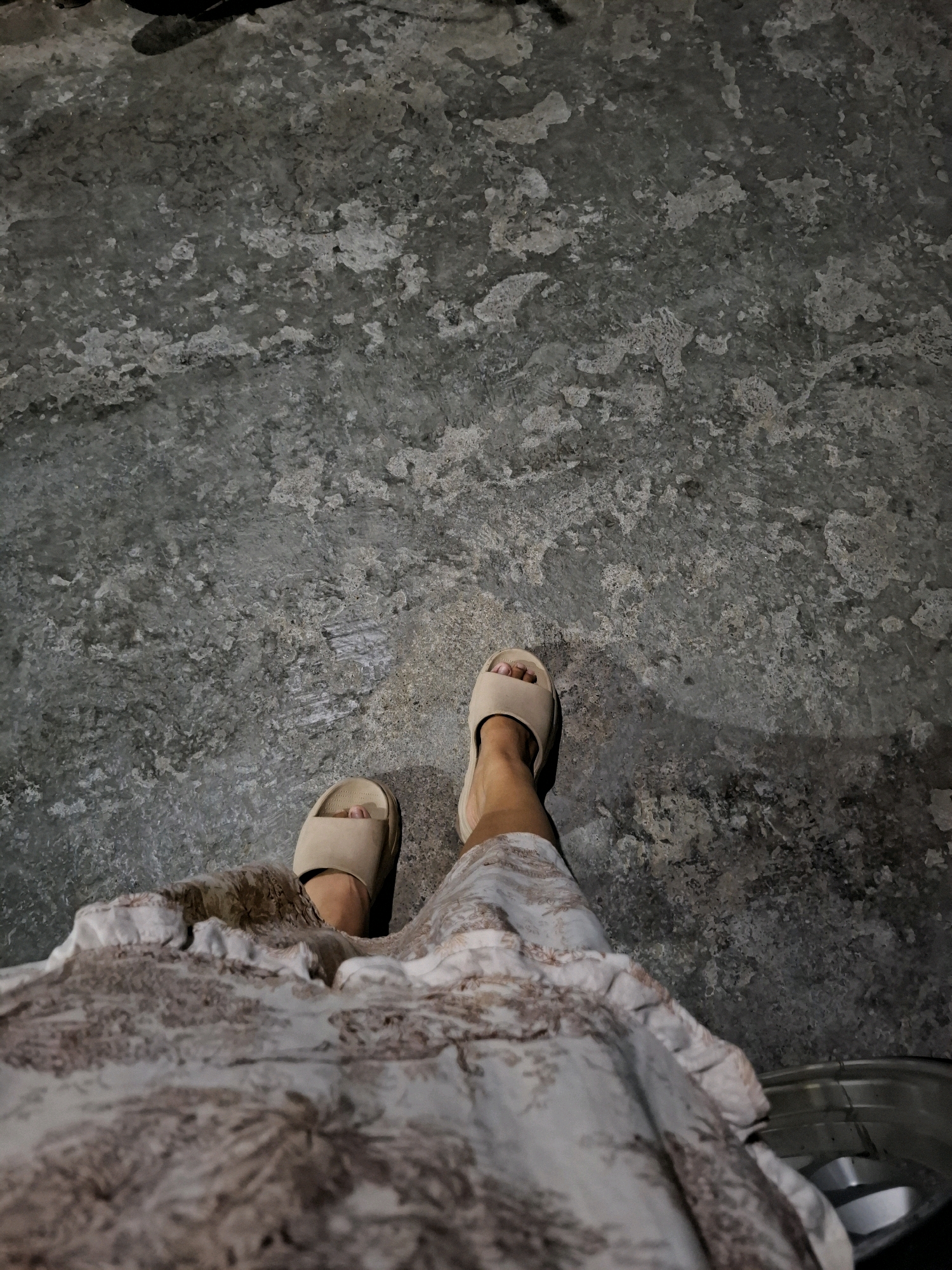 A low-angle, close-up shot depicts a person wearing light brown slides and a patterned dress standing on a textured, concrete surface. The concrete appears weathered with various lighter and darker patches, suggesting an outdoor or industrial setting. The dress has a white base with a brown, repeating pattern that seems to be floral or botanical in nature. The person's feet and the lower portion of their legs are visible, with one leg slightly in front of the other. The background is a continuation of the same concrete texture, extending upwards and out of frame. The lighting suggests it might be evening or a dimly lit area, casting subtle shadows. The image appears to be taken in Sembung, Indonesia, though no specific landmarks are identifiable in the frame.