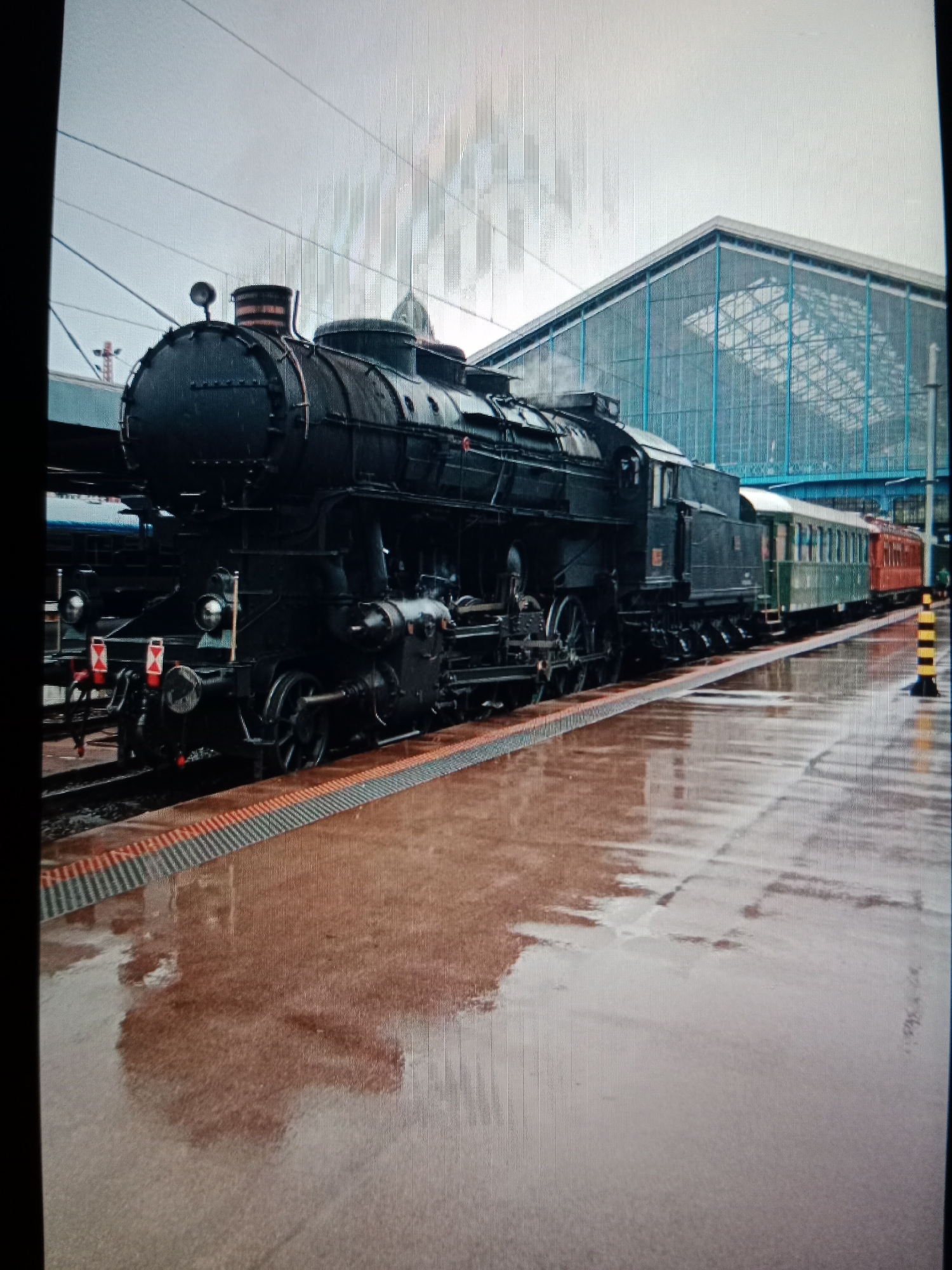 The image displays a black steam locomotive and its carriages at a train station platform on what appears to be an overcast day. The large, powerful locomotive, with its classic design, is the primary subject, positioned on the tracks and emitting smoke or steam from its chimney. Attached to it are at least two passenger carriages, one dark green and the other reddish-orange, suggesting vintage rolling stock.
The train rests beside a wide, reddish-brown platform that is visibly wet and highly reflective, mirroring the train and elements of the station structure above. In the background, a modern train station building dominates the upper right, featuring a distinctive roof and wall constructed from glass and metal, characterized by blue structural framing. The sky overhead is a uniform, light grey, indicating cloudy weather.
On the platform, near the rear of the visible carriages, a black and yellow striped bollard is present. The front of the locomotive shows some red and white markings on its buffer beam, and small, indistinct white numbers are visible on its boiler and on a red plate on its side, but specific text or numbers are not clearly legible. The scene evokes a blend of historical transportation with modern infrastructure, possibly during a special event or heritage operation, under conditions of recent rainfall.