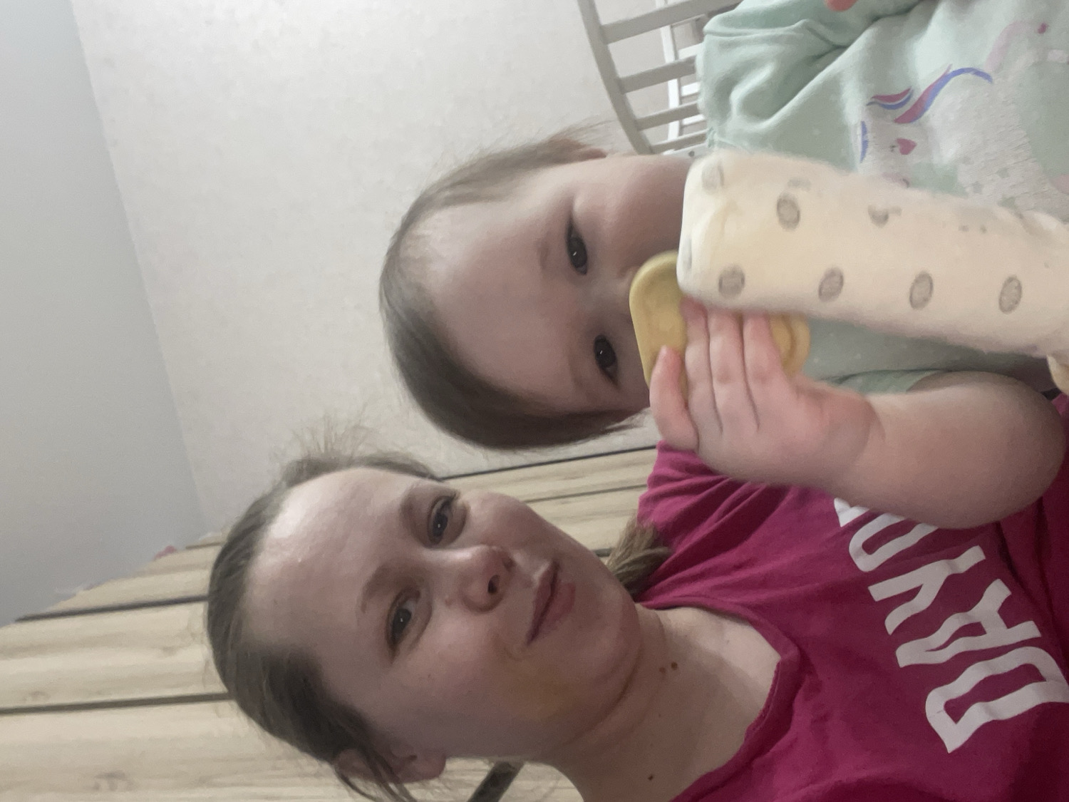 This close-up, low-angle selfie captures an adult female and a young child in an indoor setting, likely a home in Chaikovsky, Russia. The image is notably tilted, giving it a candid, personal feel.
The adult, positioned in the lower left portion of the frame, has light brown hair, pulled back but with some strands framing her face. She has fair skin and light-colored eyes, looking directly at the camera with a gentle, slight smile. She is wearing a fuchsia or magenta t-shirt, on which white capital letters spelling "DAY" are partially visible. A yellowish smudge is present on her right cheek or jawline.
The child, a baby or toddler, is positioned above and to the right of the adult, appearing to be lying down or held. They have light brown hair and dark, watchful eyes. The child is dressed in a light green or mint-colored top with a subtle, indistinct pattern. They are actively holding a baby bottle with a yellow nipple and a light-colored, dotted cover or sleeve, seemingly in the process of drinking.
The background consists of a light-colored wall or ceiling, and what appears to be light wooden paneling or floorboards, indicating a domestic environment. A white crib railing is partially visible in the upper right, reinforcing the impression of a nursery or bedroom. The lighting is soft, characteristic of indoor conditions. The overall mood conveyed is one of affection and everyday family life.