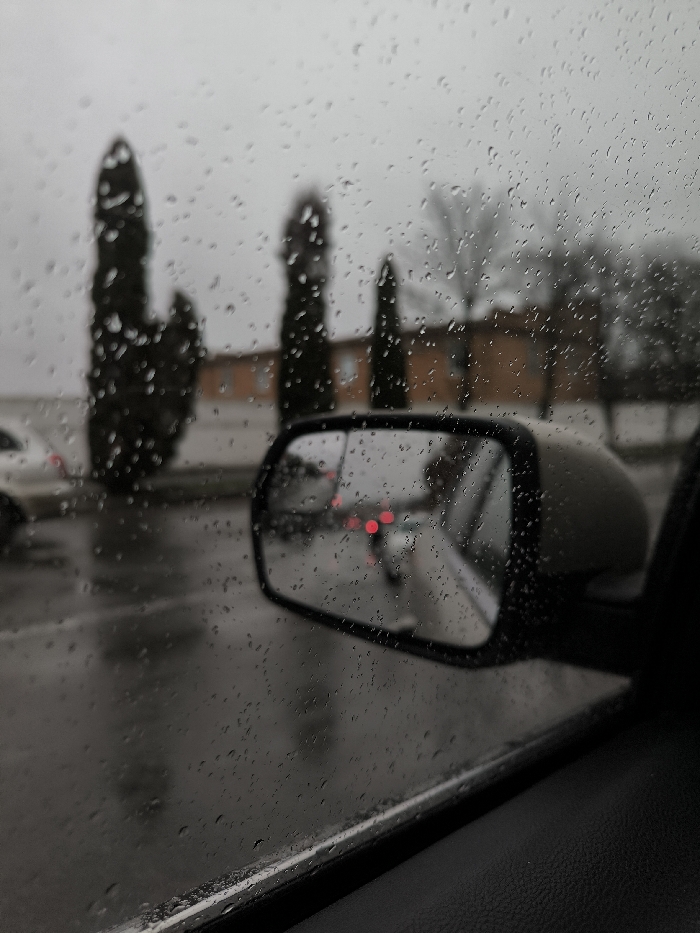 The image depicts an exterior view from inside a vehicle on an overcast, rainy day in Illintsi, Ukraine. Numerous spherical raindrops cover the car's window in the foreground, indicating active precipitation. A dark-colored car door panel or window sill is visible at the bottom right. A prominent side-view mirror, positioned on the lower-right side of the image, reflects blurred red taillights and the outline of a vehicle behind the observer's car. A faint, blurred reflection of a human figure is discernible within the mirror. Beyond the rain-streaked window and the mirror, the out-of-focus background features a wet, dark asphalt surface. Three distinct, tall, slender coniferous trees are visible on the left side, with a bare deciduous tree further right. Behind these trees, a light brown or orange building with multiple windows is present. A blurred white car is partially visible on the far left, suggesting parked or moving traffic. The sky is uniformly gray and overcast. No explicit human actions or interactions are visible.