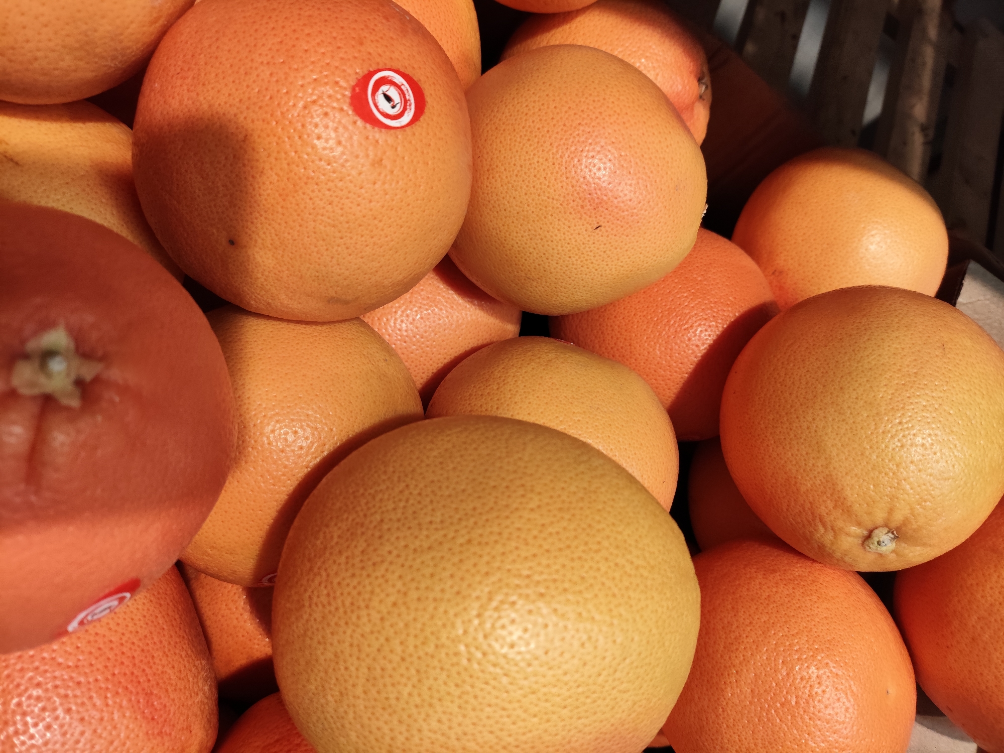 A close-up, overhead shot showcases a pile of ripe grapefruits, their textured orange rinds filling the frame. One grapefruit in the upper left section displays a small, round red and white sticker. The lighting suggests an outdoor or well-lit indoor setting, possibly a market or grocery store. The background features the indistinct slats of a wooden crate, hinting at storage or display. The image does not contain any recognizable landmarks or elements that would place it specifically in Krasnodar, Russia.