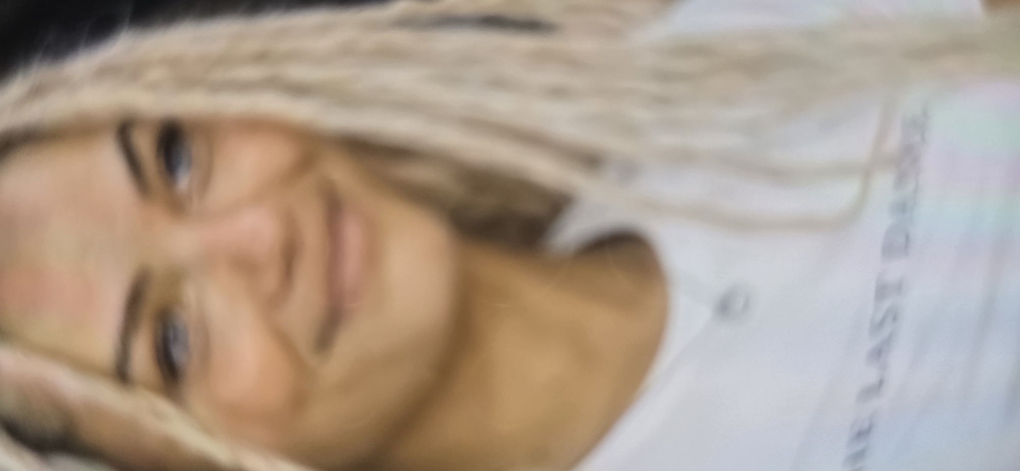 A blurred, close-up shot shows a woman with long, blonde, braided hair. She is wearing a white t-shirt with a faint necklace visible. Text is partially visible on the shirt, reading "THE LAST DANCE". The image is too blurry to determine her specific location within the city of Kurgan, Russia, or any other environmental details.