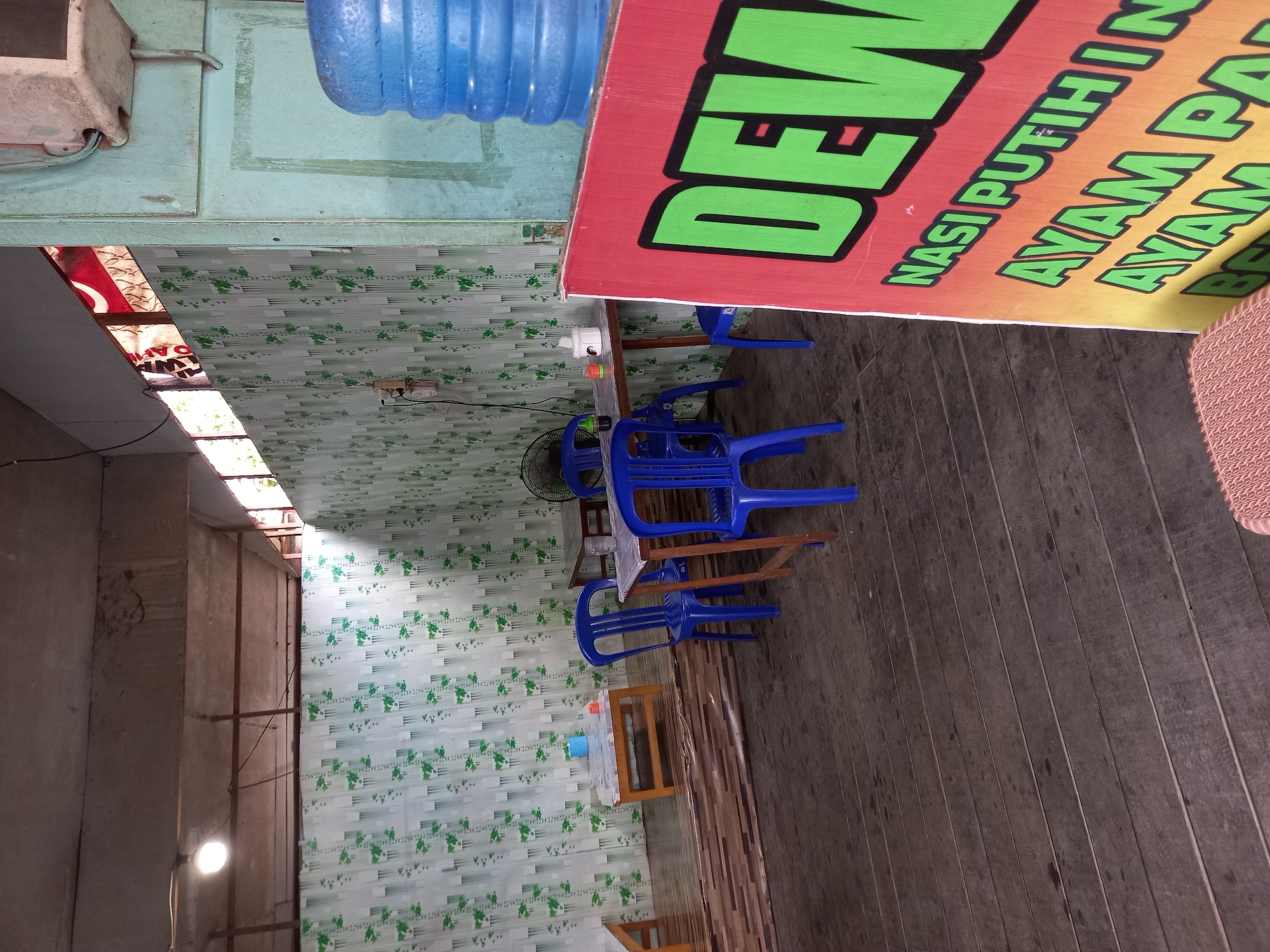 The image depicts an outdoor dining area, likely a warung or food stall, in Samarinda, Indonesia. The flooring is made of dark wooden planks, worn and showing signs of use. The walls are covered in a green and white patterned wallpaper with a leaf motif.
In the foreground, a large red banner with text is partially visible. The visible text in bright green and yellow reads "DEW" and then "NASI PUTIHIN AYAM PA AYAM". This suggests a menu offering white rice, chicken, and other dishes. To the right of the banner, a large blue water dispenser is situated.
Further back, several small wooden tables with wooden chairs, painted bright blue, are arranged. Some tables have white napkins or paper dispensers on them. A standing fan is visible behind one of the tables. The ceiling appears to be constructed from corrugated metal panels, with exposed pipes and wiring. Lighting is provided by a single bare bulb hanging from the ceiling. Natural light filters in from openings in the structure, and some windows are visible, covered by what appears to be a red and white striped fabric. The overall impression is of a functional, no-frills eatery. There are no people visible in the image.