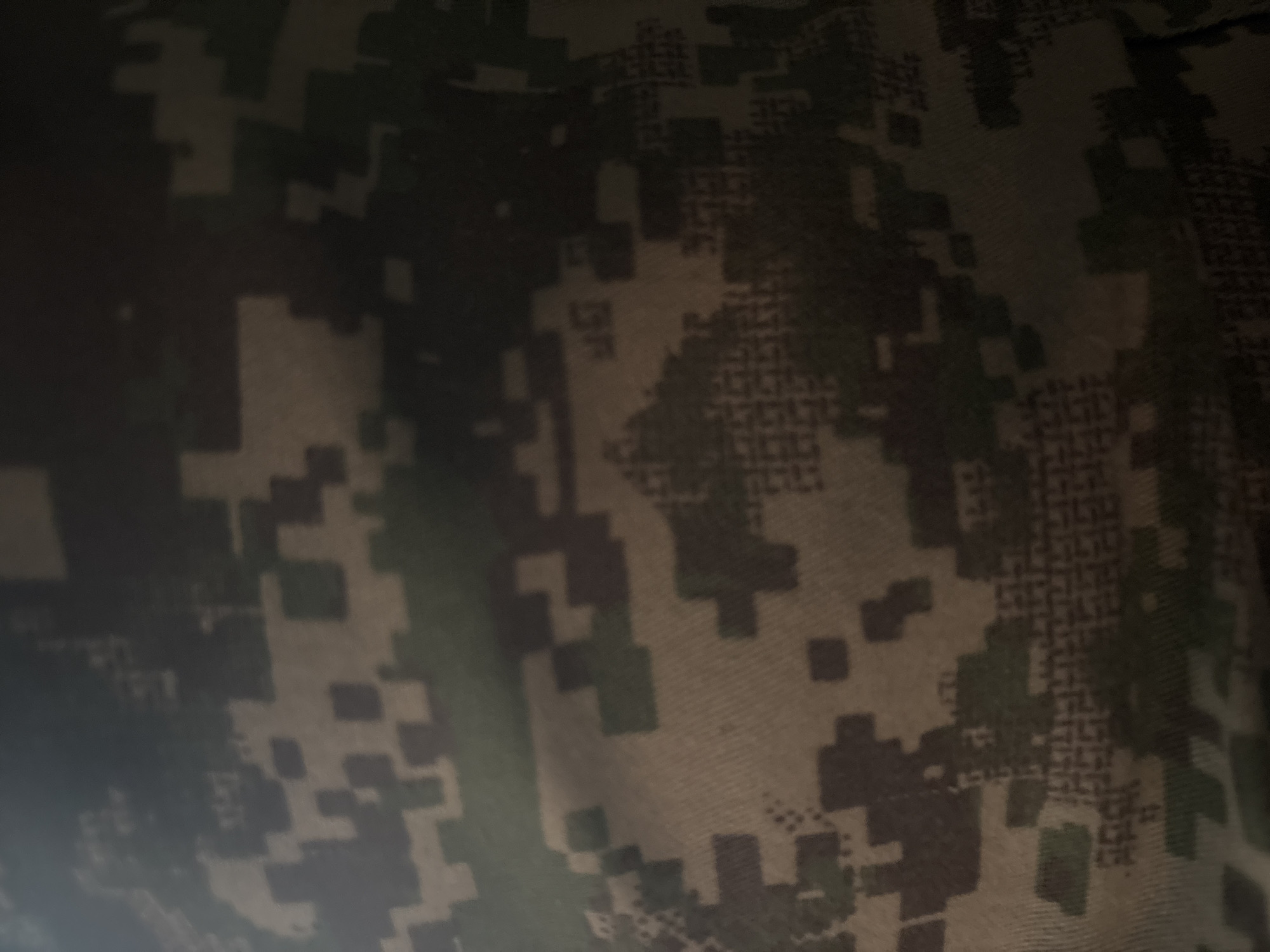 The image is a close-up, abstract shot of a digital camouflage pattern. The pattern consists of irregular, pixelated shapes in shades of brown, green, and black, designed to blend with natural surroundings. There are no discernible objects, people, actions, or interactions visible. The lighting is dim, casting shadows that obscure some of the details of the camouflage. There is no information in the image that can be used to determine its location in Ocheretianka, Ukraine, or any other specific geographical area.