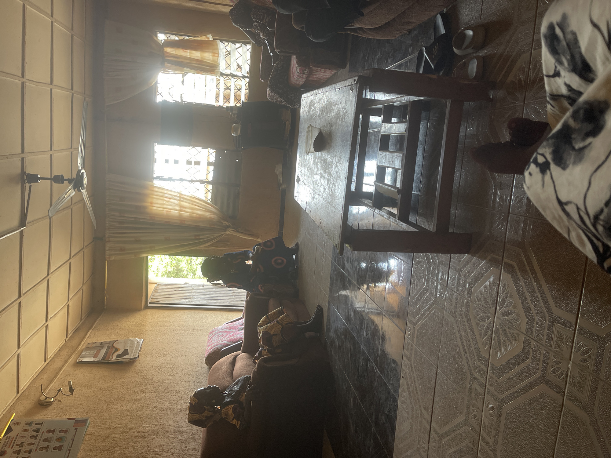 The image depicts the interior of a room in Sabon Tasha, Nigeria. The room is furnished with sofas, a wooden table with items on it, and has patterned tile flooring. Light enters through curtained windows at the top of the image and a window near the lower left. A ceiling fan is visible. People are present in the room, with one person seated on a sofa. There are also other people in the room. A newspaper and a wall lamp are seen on one wall.