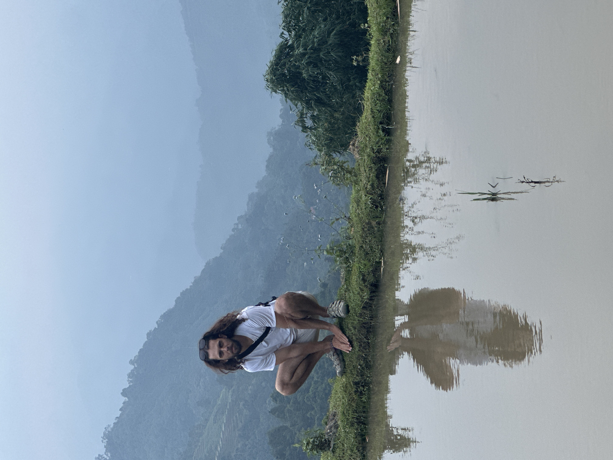 A man with long, dark hair and a beard is captured in a squatting position on the edge of a grassy bank, overlooking a calm body of water. He is wearing a white t-shirt, light-colored shorts, and grey athletic sandals. Sunglasses are perched on his head, and a dark strap, possibly from a bag, runs across his chest. He looks directly at the camera with a neutral expression, his hands resting together in front of him.
The setting is a serene, natural environment in Nậm Dịch Commune, Vietnam. To the man's right, the still water acts as a mirror, reflecting his image along with the distorted reflections of lush green trees on the opposite bank. The background features hazy, verdant mountains stretching into the distance under a soft, light blue-grey sky, indicating either an overcast day or the gentle light of dawn or dusk. The narrow strip of land where the man is positioned is covered in green grass and small plants. The scene conveys a sense of tranquility. There is no visible text in the image.