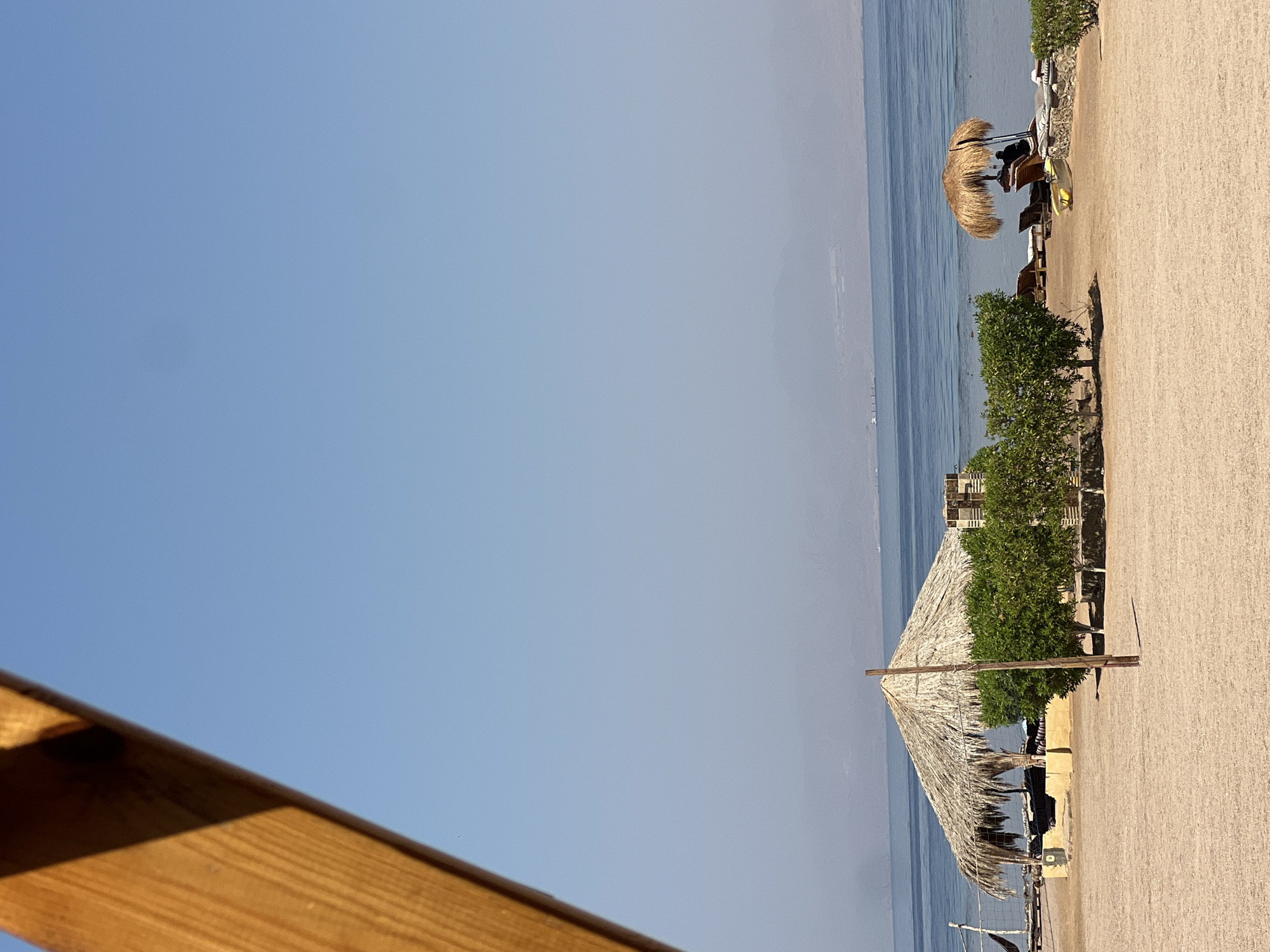 A serene beach scene unfolds under a clear blue sky, likely in Nueiba, Egypt, given the context. In the foreground, a sandy expanse stretches towards the calm, blue sea. A thatched-roof gazebo, providing shade, is nestled amongst lush green bushes. To the right, another thatched umbrella shelters a chaise lounge where a solitary figure is seated, observing the tranquil waters. The horizon is marked by distant, hazy mountains, suggesting a vast, open landscape. A volleyball net stands on the left, hinting at recreational activities. The wooden structure on the left, partially obscuring the frame, adds a rustic, tropical feel to the overall atmosphere.