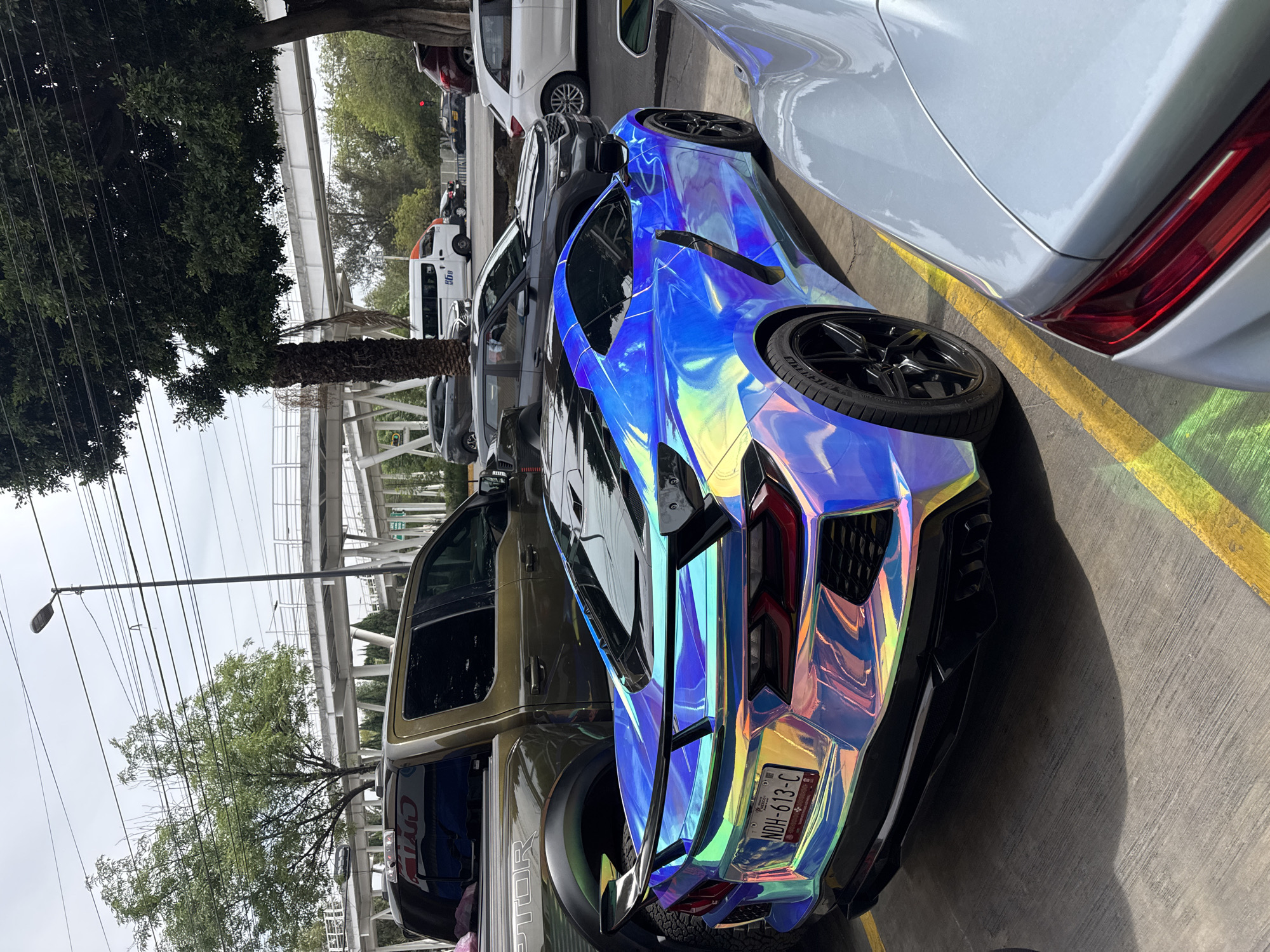 The image shows a parking lot scene in Puebla, Mexico. A brightly colored, iridescent blue and chrome car, possibly a modified Chevrolet Corvette C8, is parked between a tan Ford Raptor truck and a light silver car. The license plate of the blue car reads "NDH-613-C." The vehicles are parked on a concrete surface marked with yellow parking lines. In the background, a pedestrian bridge is visible, along with trees, power lines, and other parked cars. The sky is overcast. The photograph is taken at a low angle, emphasizing the height of the cars.