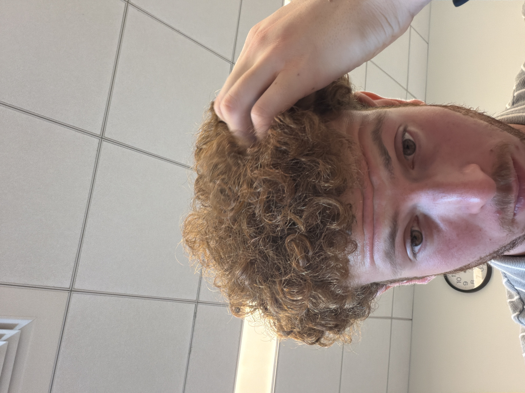 A close-up, tilted shot of a fair-skinned person with light brown, curly hair running a hand through it. The person has light eyes and a short beard. The background consists of white tiled walls and a clock. The exact location within New York City is impossible to determine from the visual information provided.