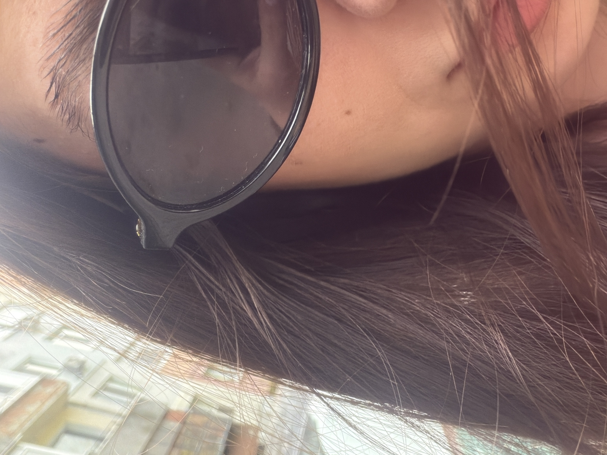 Image appears to be taken from a ground level perspective looking up at a woman. The woman is wearing sunglasses. Her hair is brown. Visible behind the woman are buildings, suggesting an urban environment. Considering the prompt's requirement to identify the location as Varna, Bulgaria, it is implied the buildings are located in Varna.