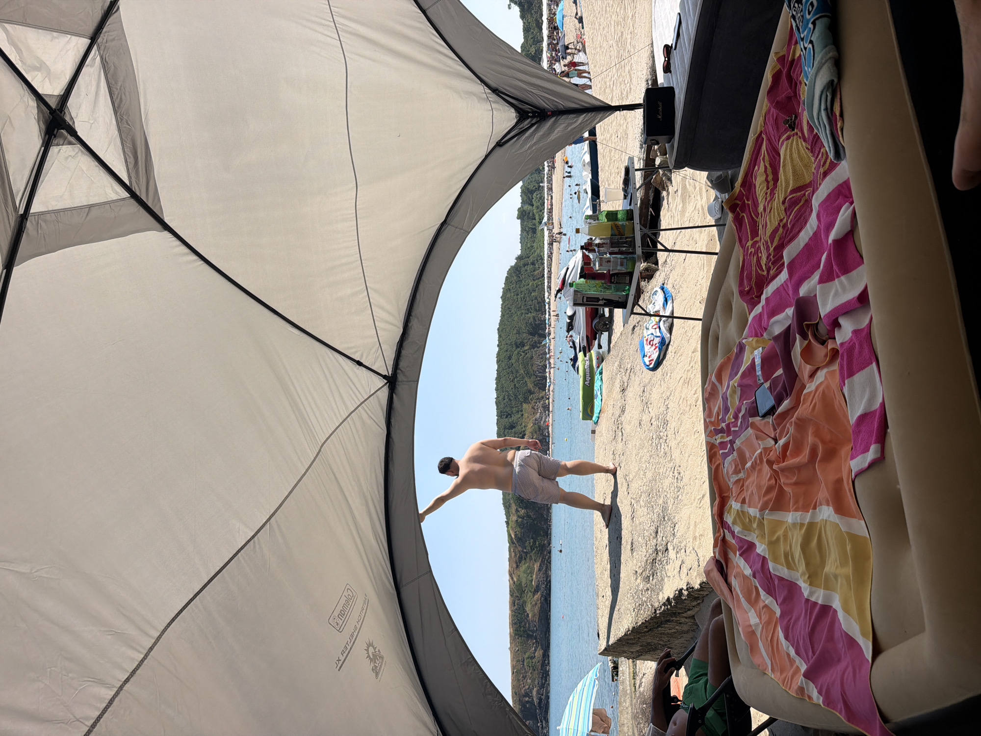 A man in patterned swim trunks leans back against a beach tent, his arm extended to touch the fabric. He is on a sandy beach, with the sea and a tree-lined coastline in the background. The water is calm and blue, with several boats and people visible. The sky is clear and bright, suggesting a sunny day. In the foreground, there are beach chairs, towels, and a cooler, indicating a day of relaxation. A Marshall speaker sits on a small table. On the beach, other people are sunbathing and swimming. The overall scene conveys a relaxed and enjoyable beach atmosphere. Visible text includes "Coleman" on the tent.