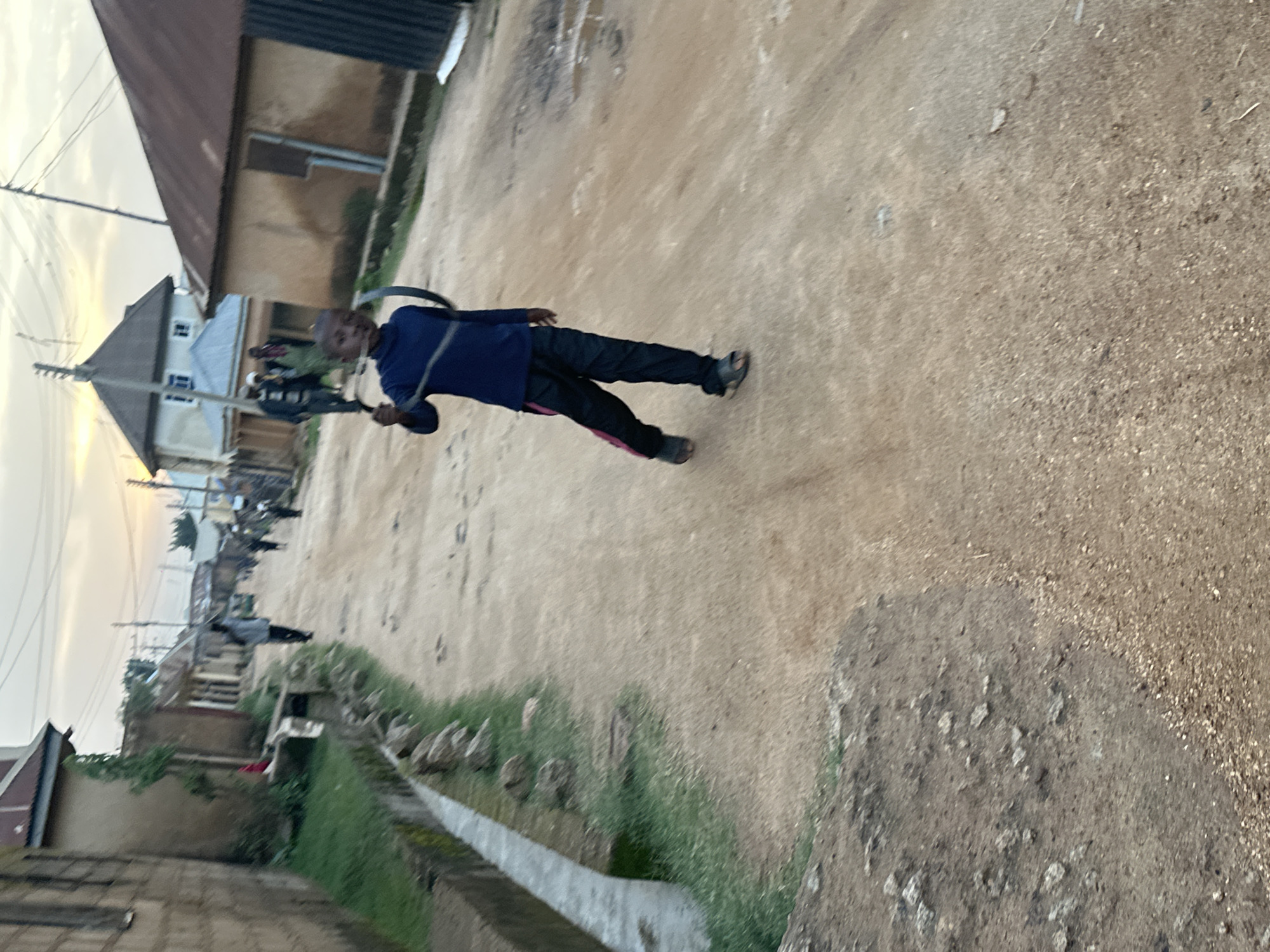 The image depicts a young boy in Gwong, Nigeria, seemingly engaged in some activity outdoors. He is dressed in a blue long-sleeved shirt and black pants with pink stripes down the side. He wears dark sandals. The boy is in motion, his body angled as if he is running or has just stopped running, with one leg extended forward. He is holding a dark object with both hands, positioned in front of his body, extending diagonally upwards. The object appears to be a stick or a similar long, thin item.
The setting is a dusty, unpaved street with buildings on either side. The structures appear to be residential, with simple architecture. One building in the foreground has a brown, corrugated metal roof. Further down the street, more houses and structures are visible. There are power lines and poles in the background, indicating the presence of electricity. A narrow, overgrown drainage ditch runs alongside the street on the right side of the image, with some rocks and vegetation visible.
The lighting suggests it is either early morning or late afternoon, with the sky appearing bright and overcast, implying a diffused light and possibly humid weather. The ground is dry and dusty, typical of a dry season or a region with unpaved roads. The overall impression is one of a typical day in a rural or semi-urban Nigerian community. The boy’s expression is not clearly discernible due to the image quality and his pose, but he appears to be focused on his activity. There is no visible text in the image.