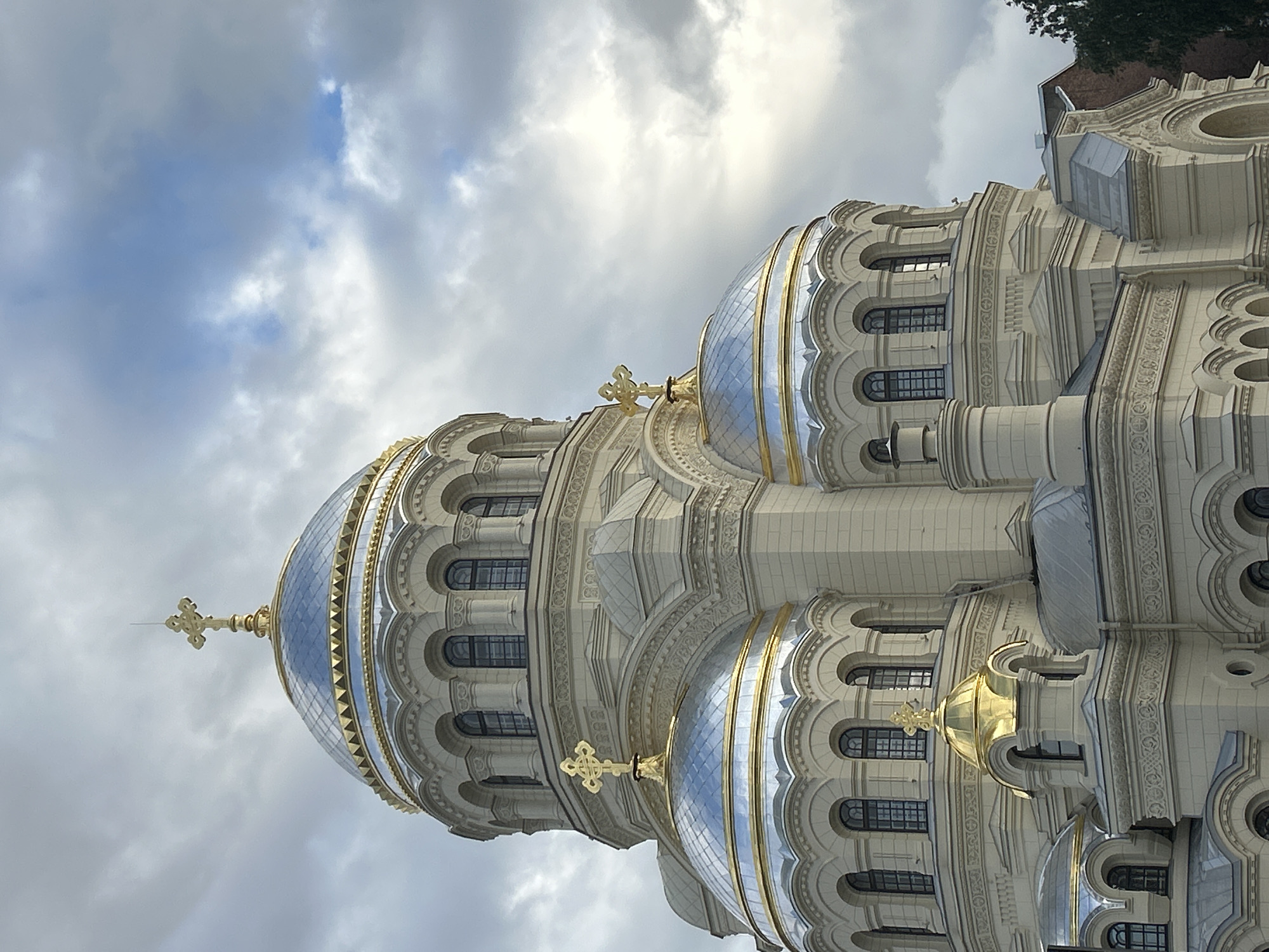 This is a low-angle shot of the upper portion of an ornate, white-and-gold domed building, likely a church or cathedral. The building's architecture is characterized by intricate carvings, arched windows, and multiple domes, some of which are a striking gold color. The larger domes are covered in reflective material, appearing silver or blue due to the sky's reflection. Golden crosses adorn the peaks of some of the domes.
The sky above is filled with dramatic clouds, ranging from bright white to darker gray, suggesting either overcast conditions or a mix of sun and clouds. The lighting indicates it is daytime, though the specific time is not clear due to the cloud cover.
No people are visible in the image. The focus is entirely on the impressive architectural details of the building. The overall mood is one of grandeur and solemnity, amplified by the cloudy sky. The image is oriented vertically, with the building's structure appearing to stretch upwards. There is no visible text within the frame.