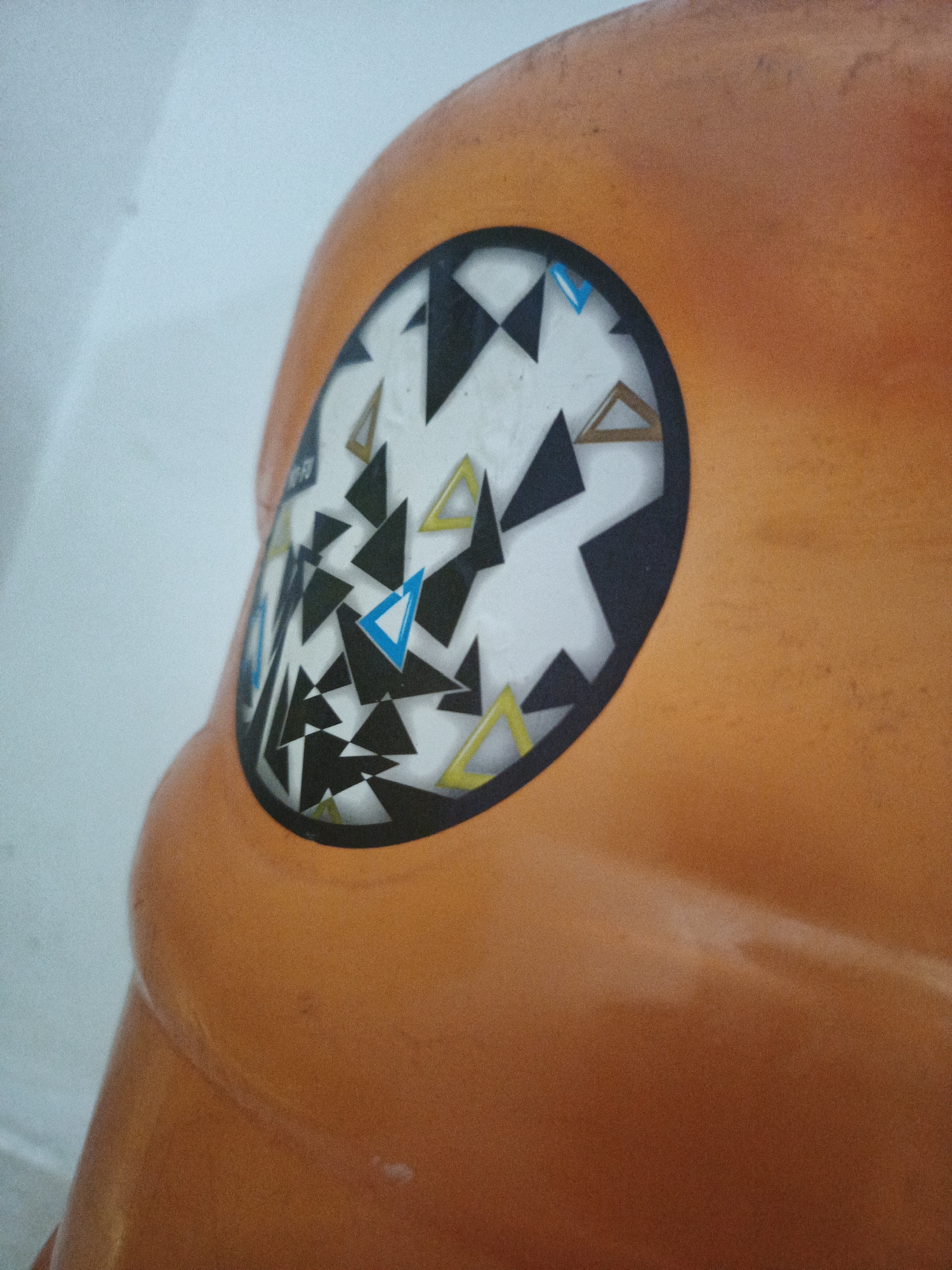The image shows a close-up of an orange plastic object, possibly a child's toy or furniture, featuring a circular sticker or design. The design consists of numerous overlapping triangles in black, white, blue, and yellow. The object's surface appears smooth and slightly reflective. The background is a plain, light-colored wall. The image does not contain information that allows for the identification of the location in Los Robles, Venezuela, as there are no identifiable landmarks or contextual clues within the frame.
