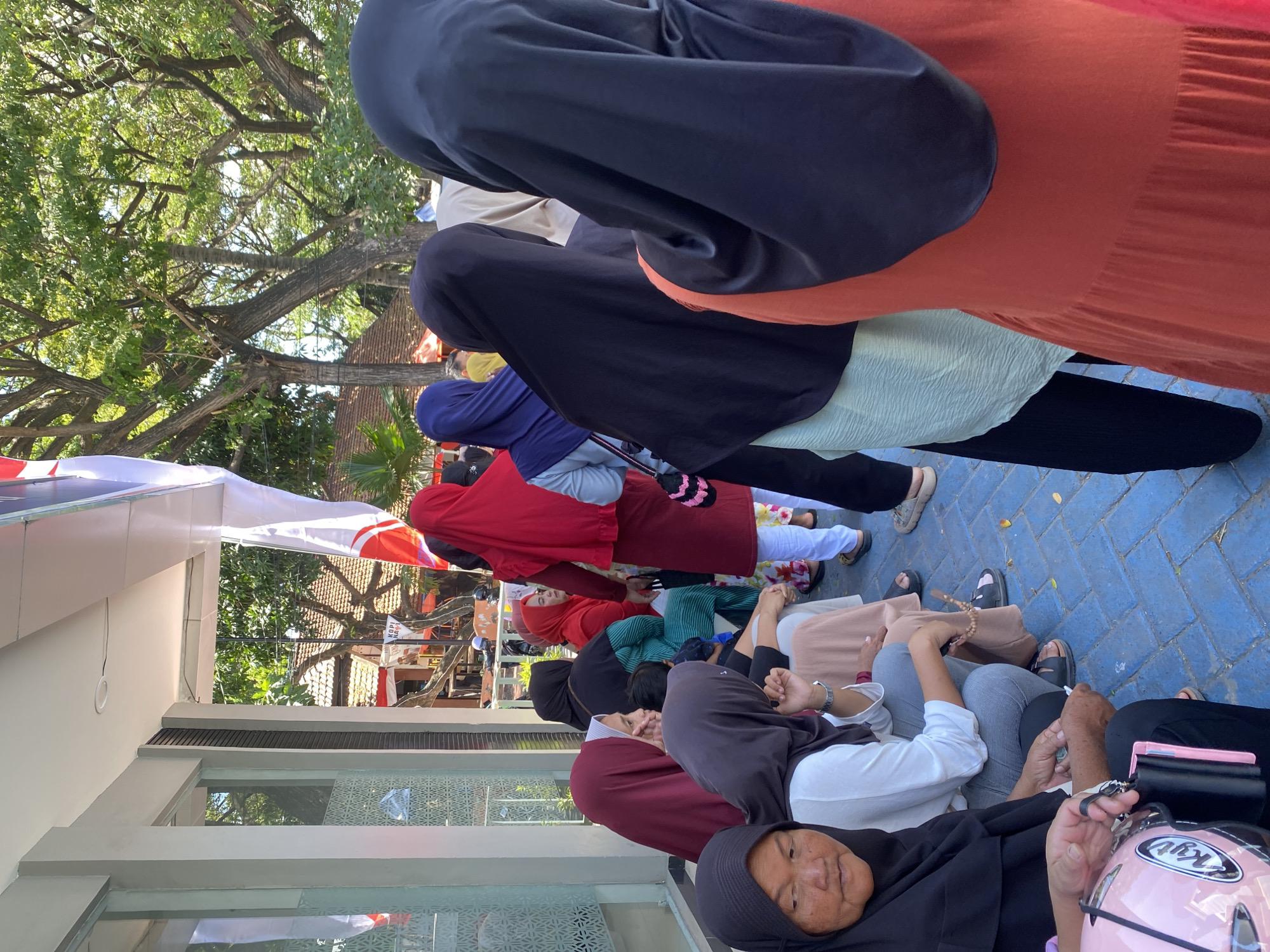 A group of people, primarily women wearing hijabs and long dresses or shirts, are gathered outdoors, sitting and standing in front of a building. The building has a light-colored facade with glass windows and a white awning. In the background, there are trees with dense green foliage, and a red-tiled roof is visible. The ground is paved with blue and white square tiles. Some of the people are looking in different directions, while others appear to be engaged in conversation. A pink motorcycle helmet and a pink wallet are visible in the foreground. The scene suggests a public gathering or waiting area, possibly near a business or community center in Kelurahan Tappanjeng, Indonesia, given the presence of trees and architectural style.