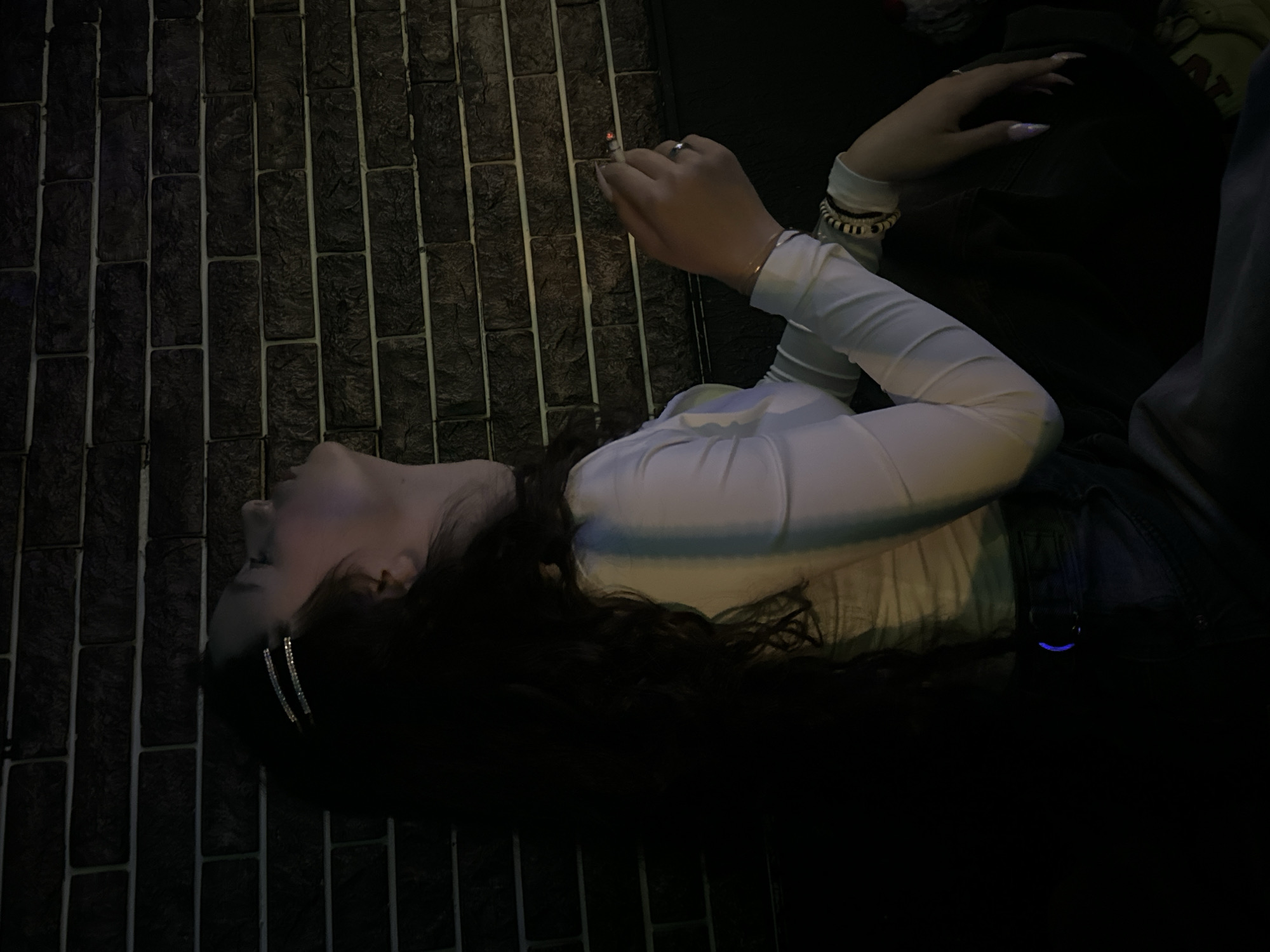 A woman with long, dark hair is lying on a textured, brick-like surface. She is wearing a white, long-sleeved top and denim overalls. Her head is tilted back, and she has two silver hair clips in her hair. In her hand, she holds a lit cigarette. The overall lighting is dim, with a focus on the subject. The scene appears to be outdoors. It is not possible to verify the location as Yerevan, Armenia, from the image alone.