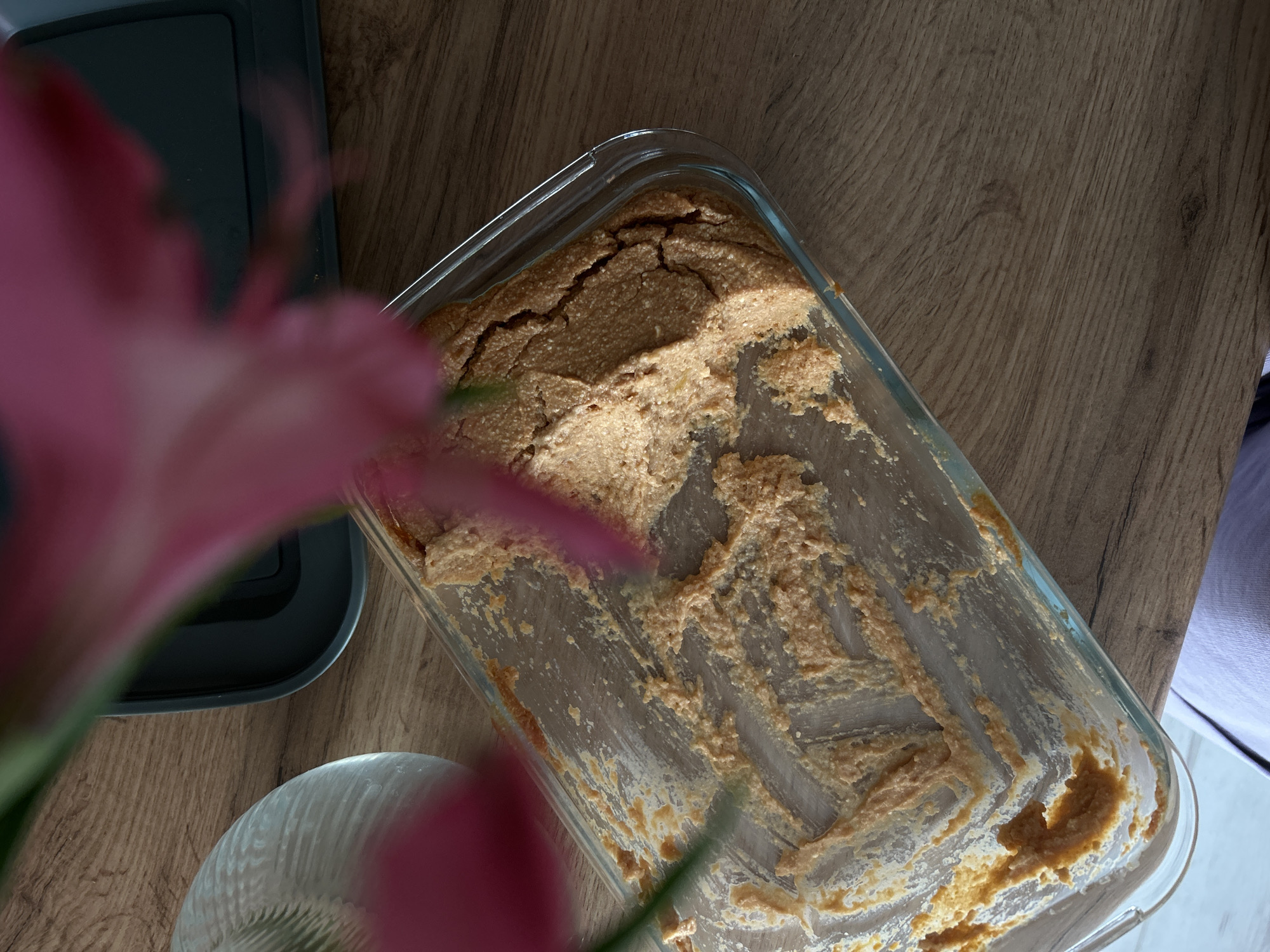 An overhead shot captures the remnants of a baked dessert in a clear glass rectangular baking dish. The dish is resting on a wooden table. The dessert, a light brown color, appears to have been mostly consumed, with only a thin layer and smears of crumbs remaining on the sides and bottom of the dish.
In the foreground and left side of the image, a large, blurred pink flower with green stems partially obstructs the view of a dark gray rectangular object, possibly a tray or lid. To the bottom left of the baking dish, a portion of a clear, ridged glass bowl is visible, also out of focus.
The lighting suggests it is daytime, with sunlight casting a warm glow on the wooden surface, highlighting the wood grain. The overall scene evokes a sense of post-meal tidiness, with the dessert dish waiting to be cleaned. No text is visible in the image.