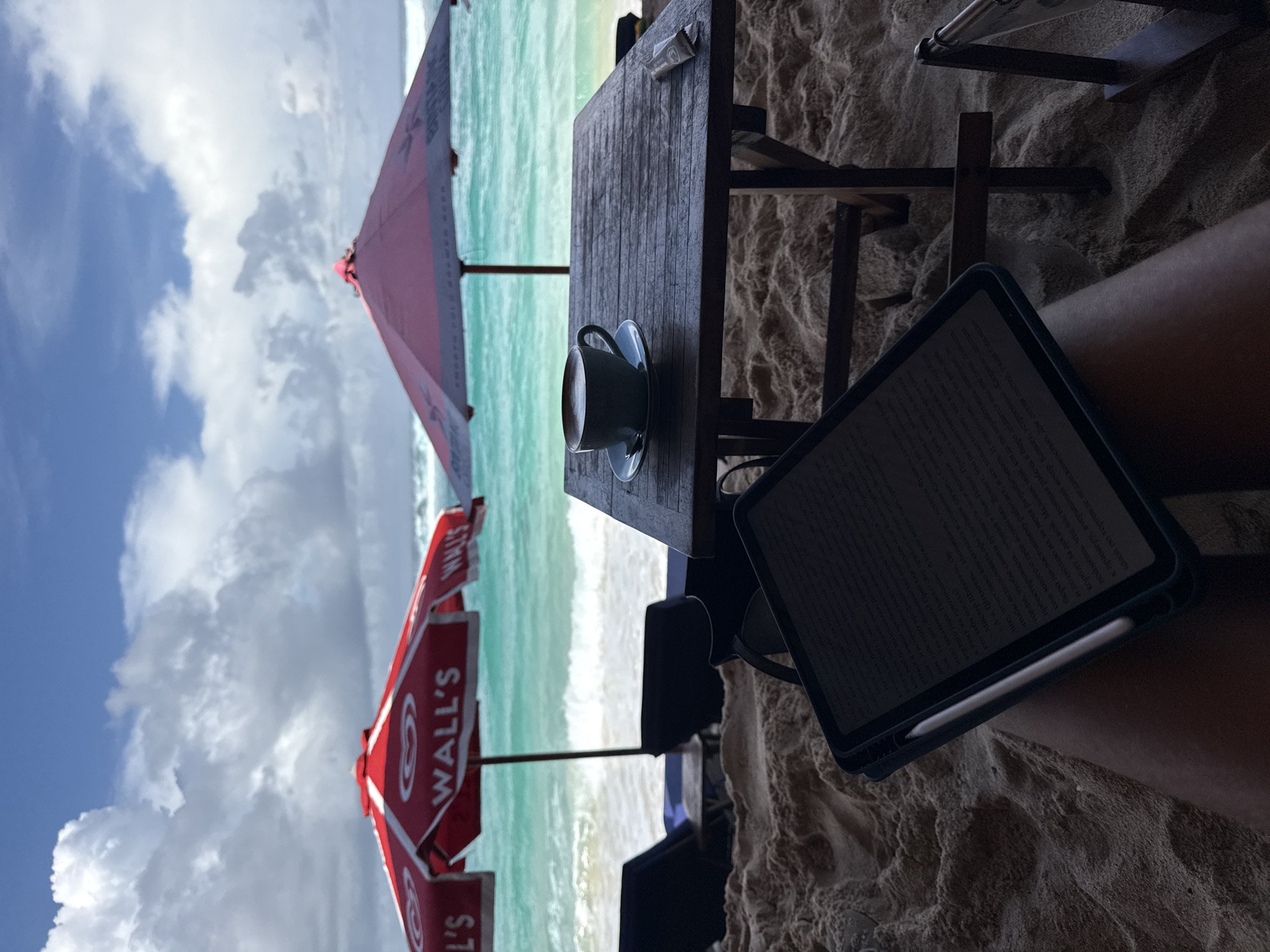 This image depicts a relaxing beach scene, likely from a first-person perspective, looking out towards the ocean on a bright, sunny day in Kuta Selatan, Indonesia.
In the immediate foreground, the viewer's legs are visible, resting on the sand. An open tablet, appearing to be an iPad, is propped on the lap, displaying an e-book or document. An Apple Pencil is magnetically attached to the side of the tablet. The screen shows dense text primarily in Russian Cyrillic script, with phrases like "Да будет тебе отлично..." and "Мистер Штенгель..." clearly discernible, indicating a narrative or descriptive content.
A rustic wooden table and chair set are positioned on the sand. On the table, a cup with a saucer holds a beverage, and a small sachet rests nearby. Further out, several large red beach umbrellas are visible. These umbrellas prominently feature the "WALL'S" brand logo, which includes a stylized heart icon, suggesting a refreshment or ice cream vendor nearby.
Beyond the beach furniture, the vibrant turquoise and green waters of the ocean stretch into the background, with gentle white waves breaking on the shore. Above the ocean, a clear blue sky is dotted with soft white clouds, enhancing the serene and leisurely atmosphere of the scene. The overall impression is one of peaceful enjoyment and relaxation by the sea.